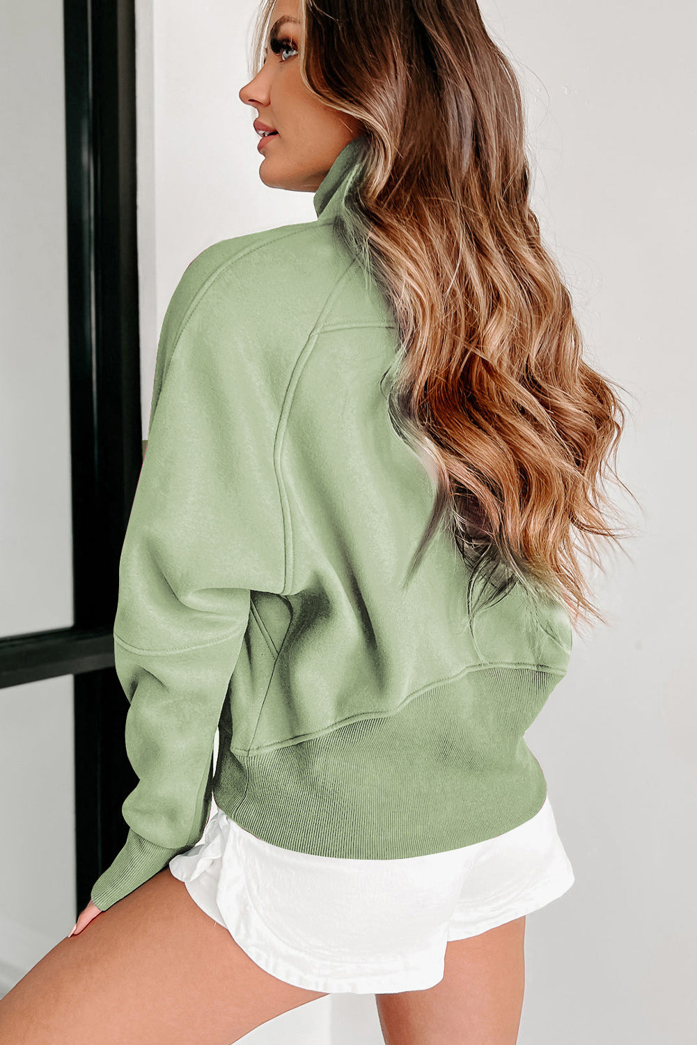 A woman with long, wavy hair wears a versatile Shewin Gray Zip Up Stand Collar Ribbed Thumbhole Sleeve Sweatshirt and white shorts, standing with her back partially turned.