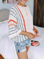 A woman wearing the Rosa Clothing Boho Flared Sleeve Loose Knit Sweater and denim shorts stands in a bedroom, with a bed and a plate of donuts visible in the background.
