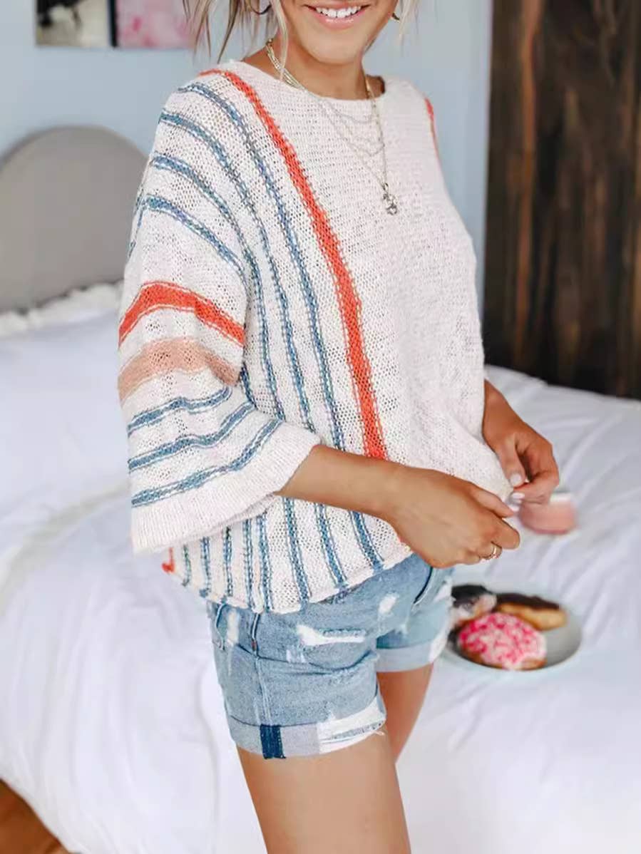A woman wearing the Rosa Clothing Boho Flared Sleeve Loose Knit Sweater and denim shorts stands in a bedroom, with a bed and a plate of donuts visible in the background.