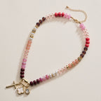 The Textured Glass Bead Necklace with Charm Trio by Lilla Haven features pink, purple, and gold beads accented with gold-toned spacers, and showcases a gold cross, a clover charm, a rectangular gem charm, all finished with a gold chain extender.