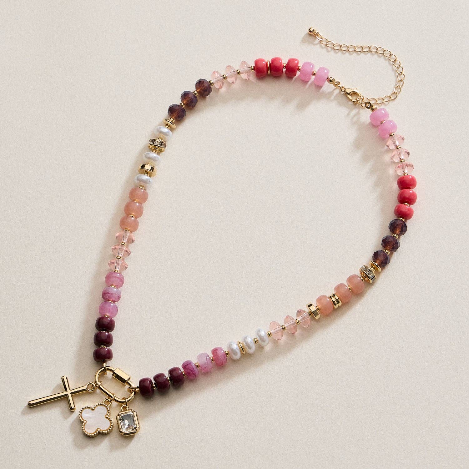 The Textured Glass Bead Necklace with Charm Trio by Lilla Haven features pink, purple, and gold beads accented with gold-toned spacers, and showcases a gold cross, a clover charm, a rectangular gem charm, all finished with a gold chain extender.