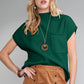 A woman stands against a gray background, wearing the Shewin Patch Pocket Ribbed Knit Short Sleeve Sweater in green, along with a necklace and a brown button-up skirt. This ensemble is ideal for transitional weather, offering versatile knit fabric that combines style and comfort.