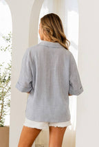 A woman with long hair faces away indoors by arched openings, wearing the Sew In Love Relaxed Fit Collared V Notch Neck Striped Top and white frayed shorts.