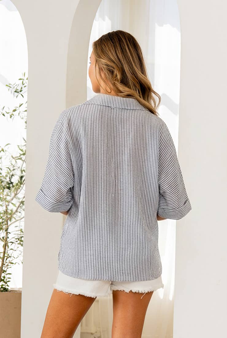 A woman with long hair faces away indoors by arched openings, wearing the Sew In Love Relaxed Fit Collared V Notch Neck Striped Top and white frayed shorts.