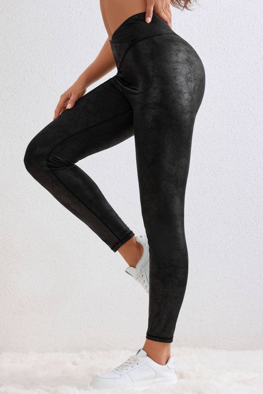 A person wears LA Society’s Black Crossed Dip Waist Sleek Leather Leggings with a unique crossed waistband and white sneakers, standing on a soft white floor against a light-colored wall.