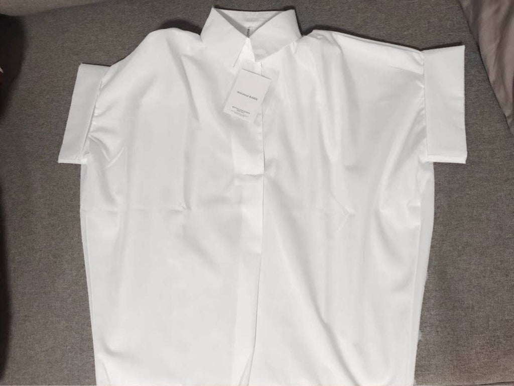 A White Relaxed Fit Short Sleeve Blouse from FASHION GO is laid flat on a grey surface, highlighting its crisp button-up design and casual silhouette, with a tag attached.
