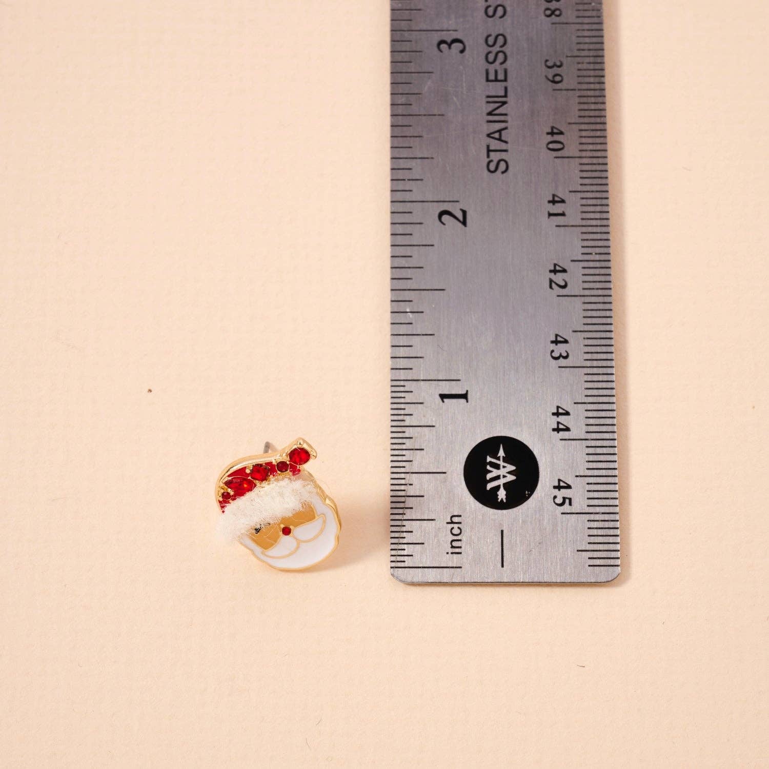 A small Santa Claus pin with red accents is placed next to a stainless steel ruler, showing its size at about 1 inch tall—perfect for pairing with the Lilla Haven Santa Claus Stud Earrings | Red Rhinestone Christmas Earrings or other festive holiday jewelry.
