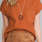 A person models the Shewin Patch Pocket Ribbed Knit Short Sleeve Sweater, a rust-colored top paired with a round pendant necklace. They complete the look with a brown high-waisted skirt featuring black buttons, perfect for transitional weather.