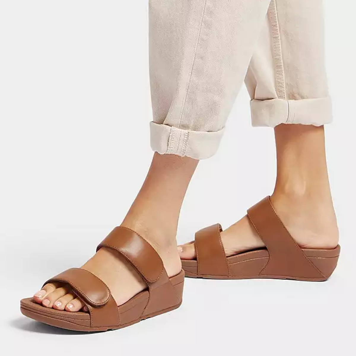 A person showcases rolled-up beige pants and minimalist-chic Lulu Adjustable Leather Slide Sandals in Light Tan by Fit Flops, featuring thick straps and possibly Microwobbleboard™ midsoles for added comfort, all set against a white background.