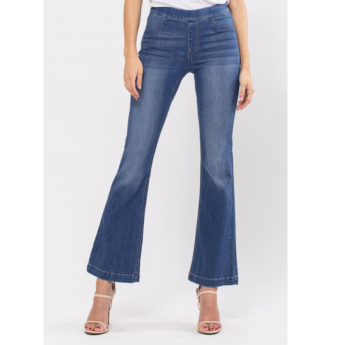 A woman stands against a plain white background wearing FASHION GO Cello Pull on Mid Rise Flare Jeans, 30" inseam in Medium Wash, paired with beige open-toe heels.