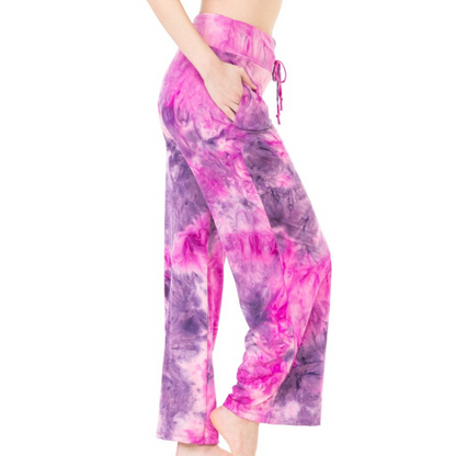 A person is wearing pink and purple tie-dye wide-leg pants with hands in the functional pockets. The image shows the FASHION GO Buttery Soft Pajama Pants with Drawstring from the side, highlighting their comfort and style.