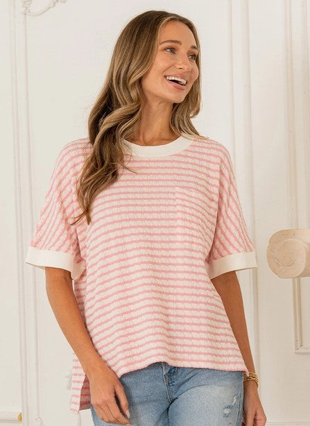A woman stands indoors, smiling, wearing the Sew In Love Relaxed Fit Cuffed Sleeves Striped Knit Top in light pink and white stripes with white trim, paired with light blue jeans.