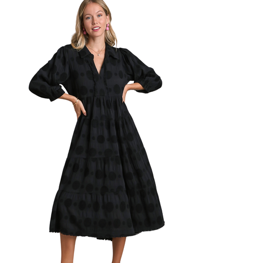 A woman stands confidently with hands on hips in the Textured Dot Tiered Midi Dress in Black by Umgee—a black, long-sleeved, tiered dress featuring subtle polka dots—set against a white background.