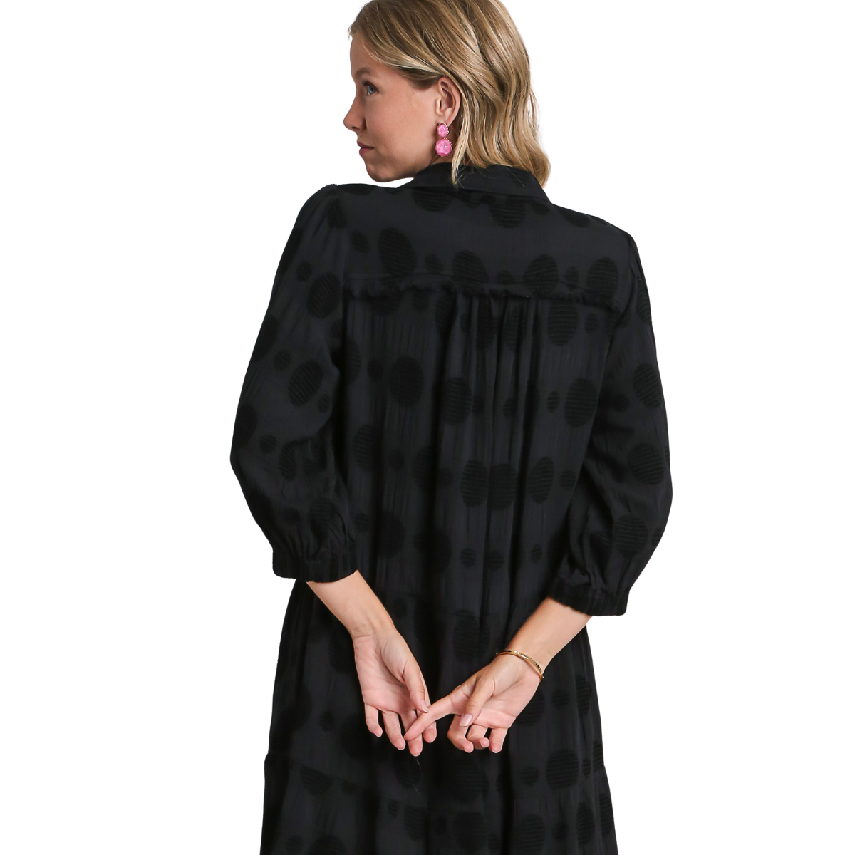 A woman stands with her back to the camera, hands clasped, wearing the Textured Dot Tiered Midi Dress in Black by Umgee and pink earrings against a white background.