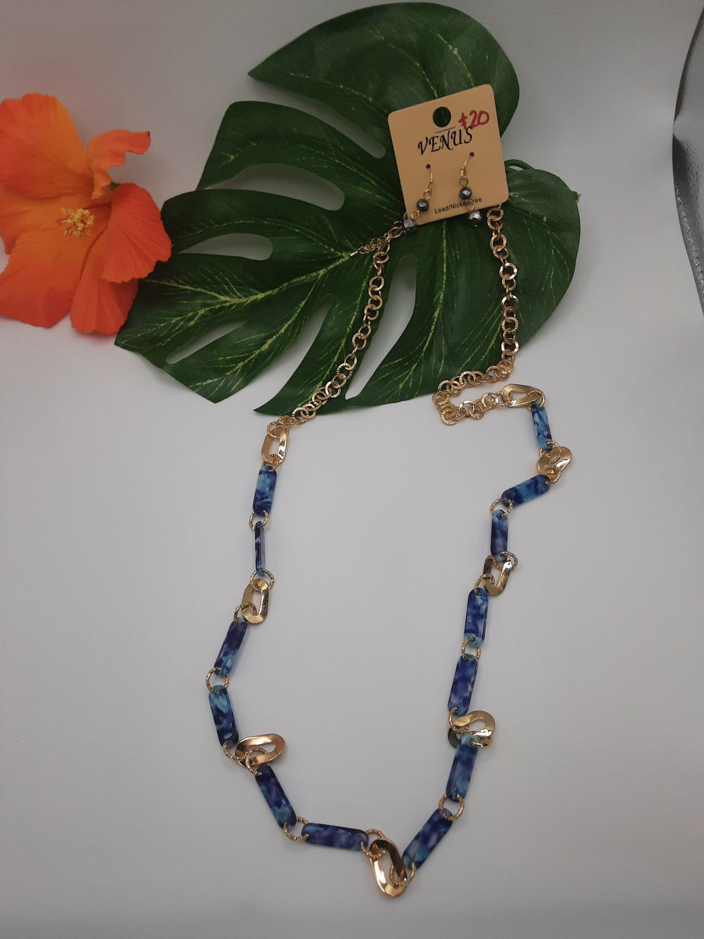 The VENUS NE6996BU BLUE chain necklace by SPECIAL EFFECTS is displayed with matching earrings on a green leaf background, accompanied by an orange flower on the side.