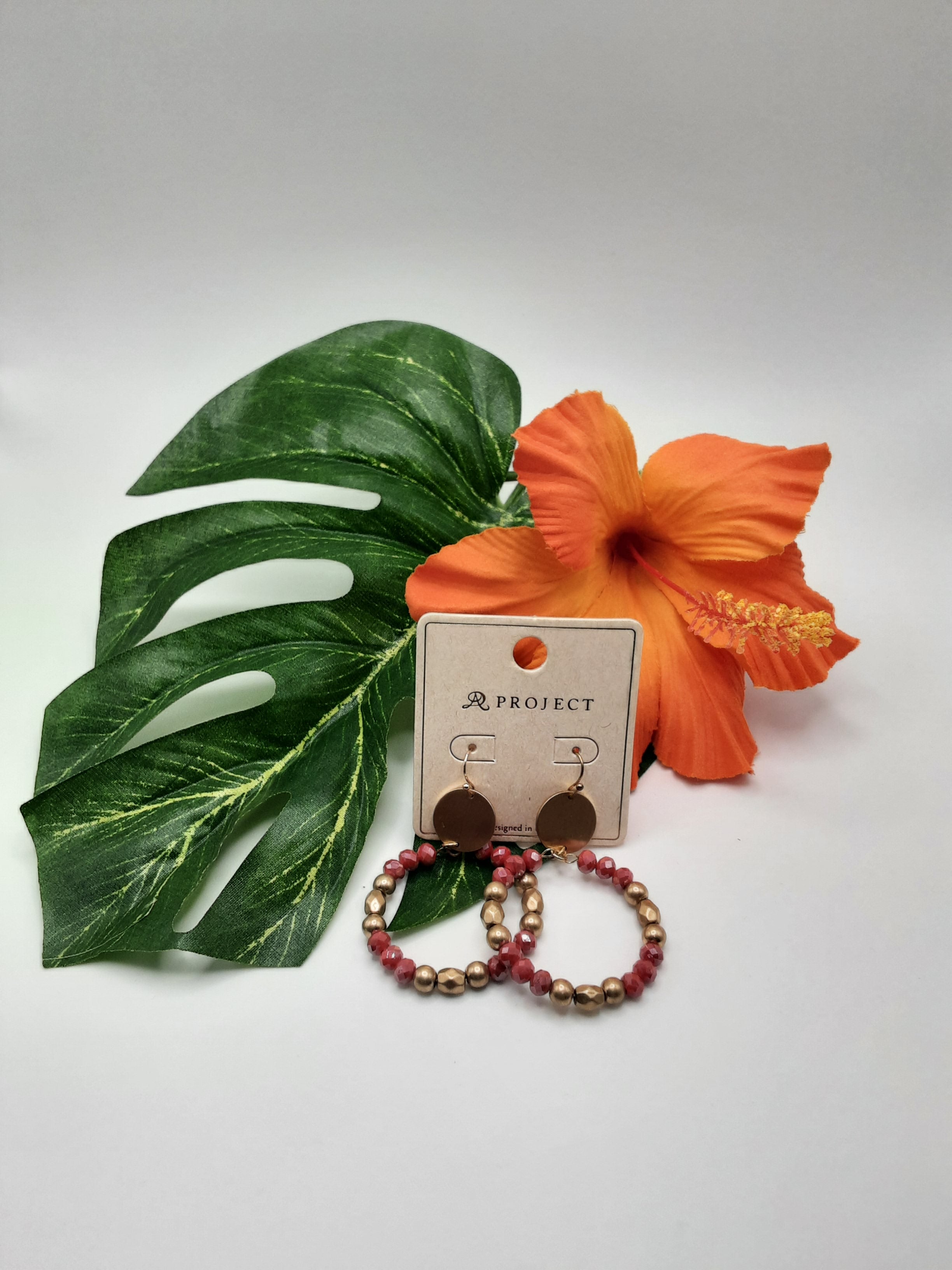 The APROJECT QE2264 EARRINGS by SPECIAL EFFECTS, featuring a pair of gold disc earrings and a set of gold and red beaded hoop earrings, are beautifully displayed alongside a green leaf and an orange hibiscus flower. Main image