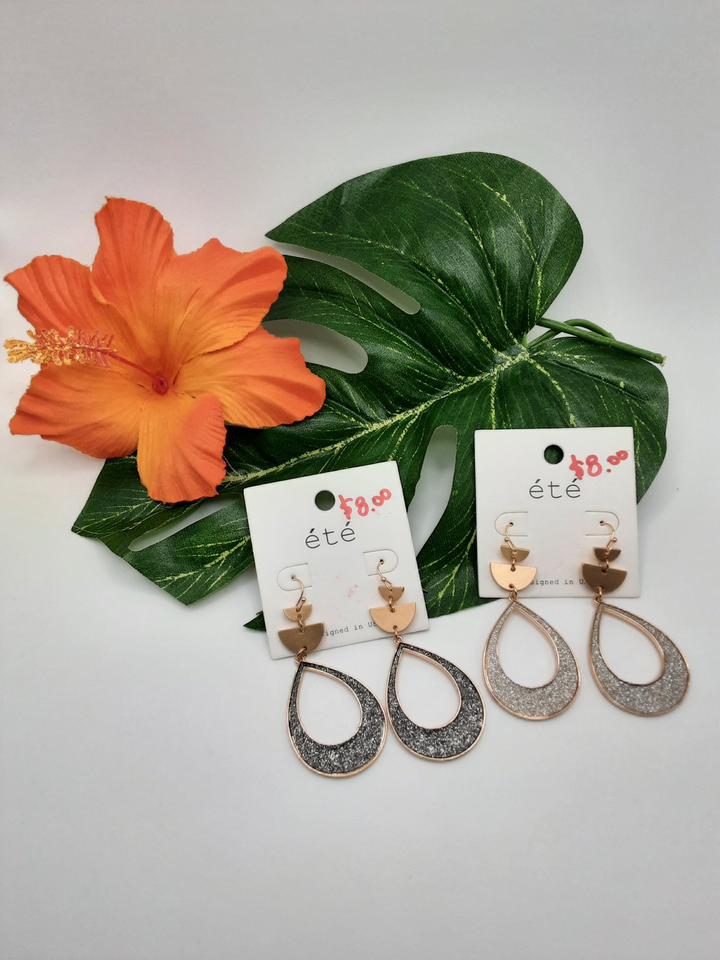 Two pairs of the SPECIAL EFFECTS GLITTER TEARDROP EARRING, boasting a silver glittery teardrop design and gold butterfly accents, elegantly displayed on white cards amidst a large green leaf and an orange hibiscus flower.