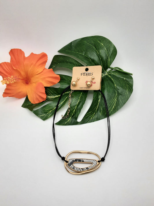 The VENUS NE5940GD SNAKE necklace by SPECIAL EFFECTS, featuring an abstract gold and silver pendant, is displayed on a leafy background beside an orange hibiscus flower. A pair of matching gold and pearl earrings are attached to the necklace card.