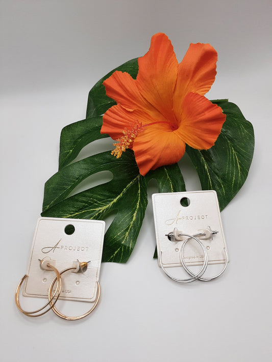 Two pairs of APROJECT KE2165 HOOPS by SPECIAL EFFECTS, one gold and one silver, are displayed on white cards alongside a bright orange hibiscus flower and green leaves.