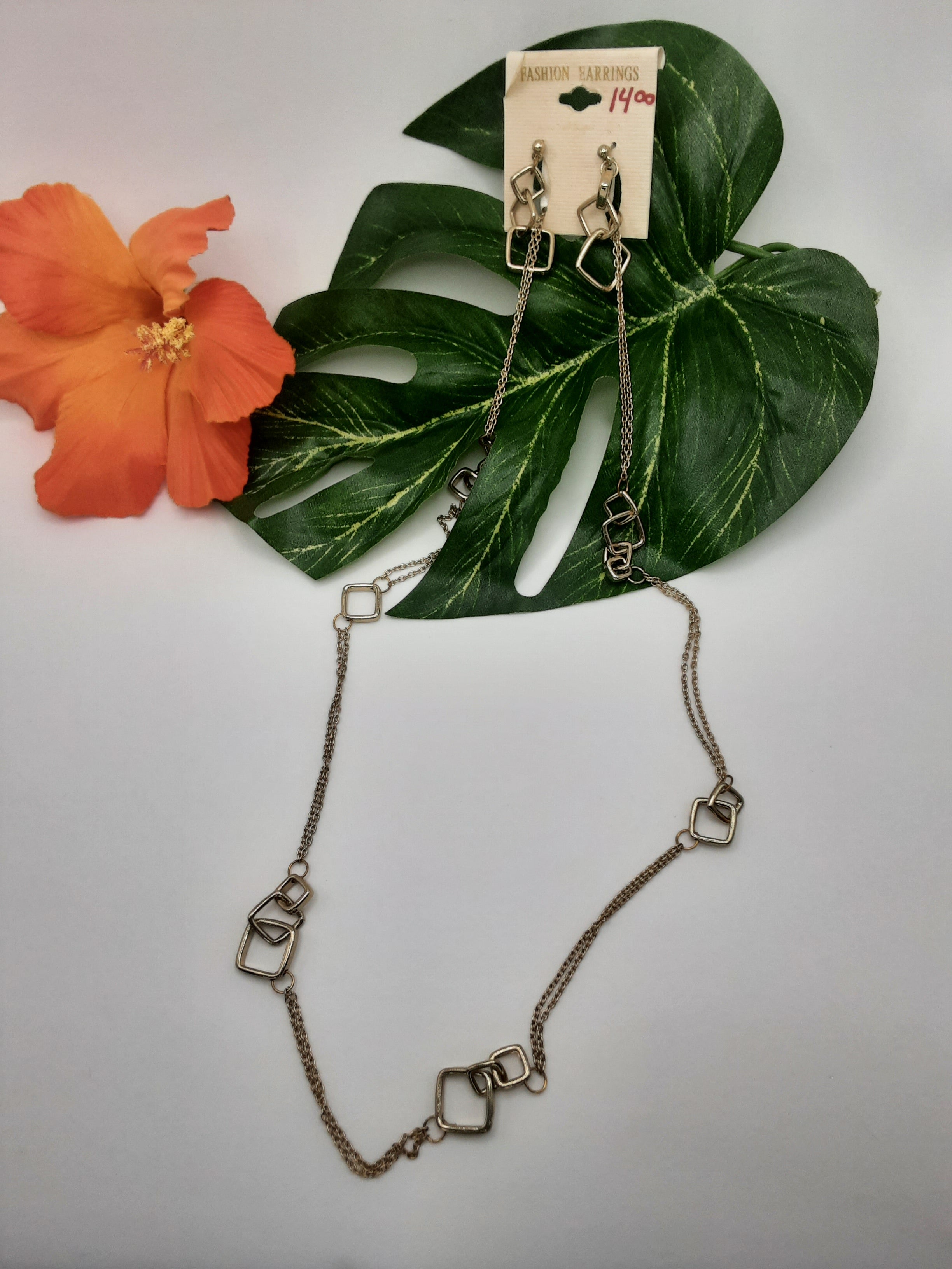 A pair of SPECIAL EFFECTS FASHION 15407 GOLD earrings on a display card priced at $14.00, accompanied by a matching necklace with geometric links, set against green leaves and an orange flower. Main image