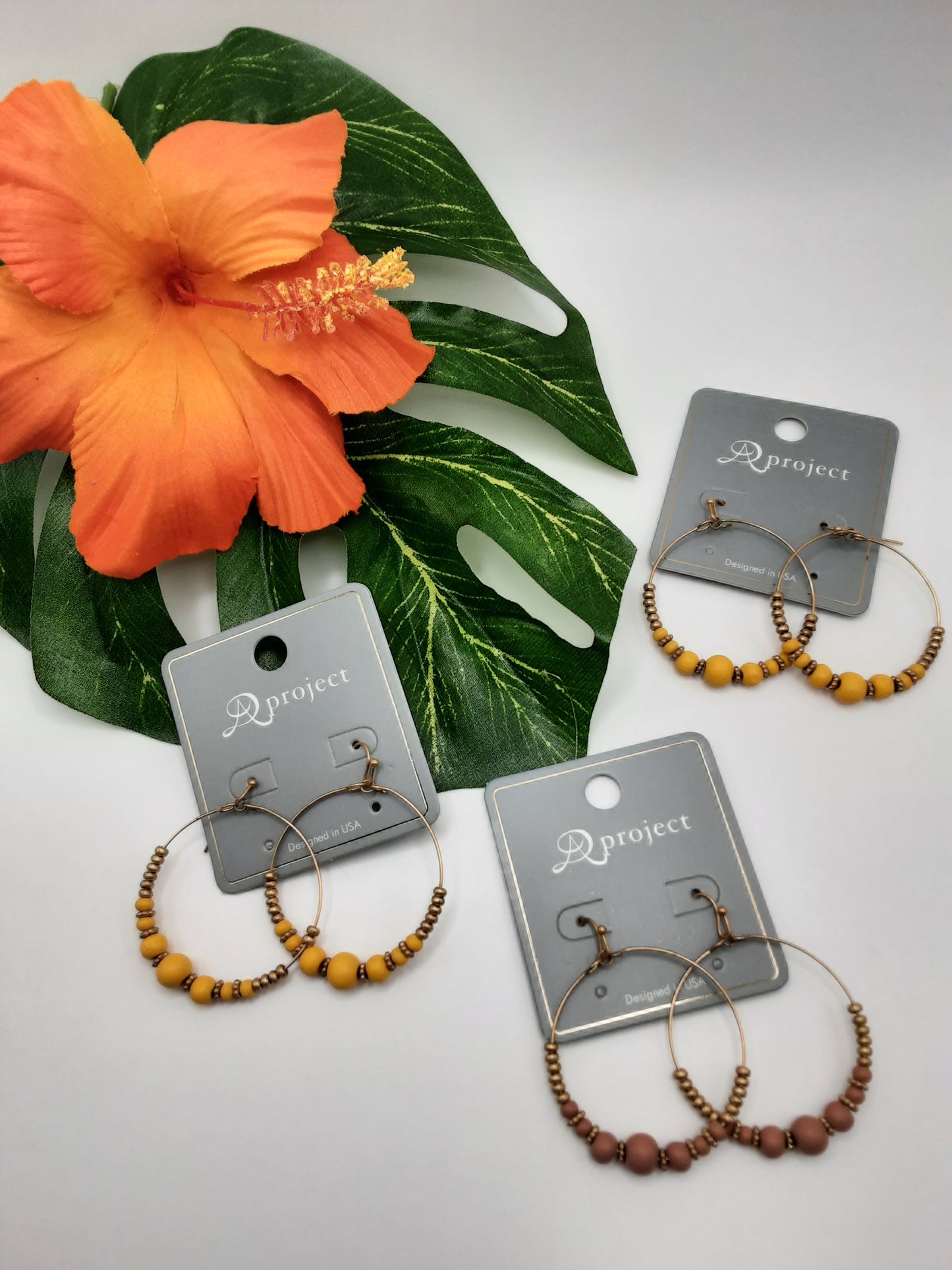 Three pairs of beaded hoop earrings from the SPECIAL EFFECTS collection, A PROJECT 3844 WOOD, are displayed on paper cards. An orange hibiscus and a green leaf are also visible in the background.