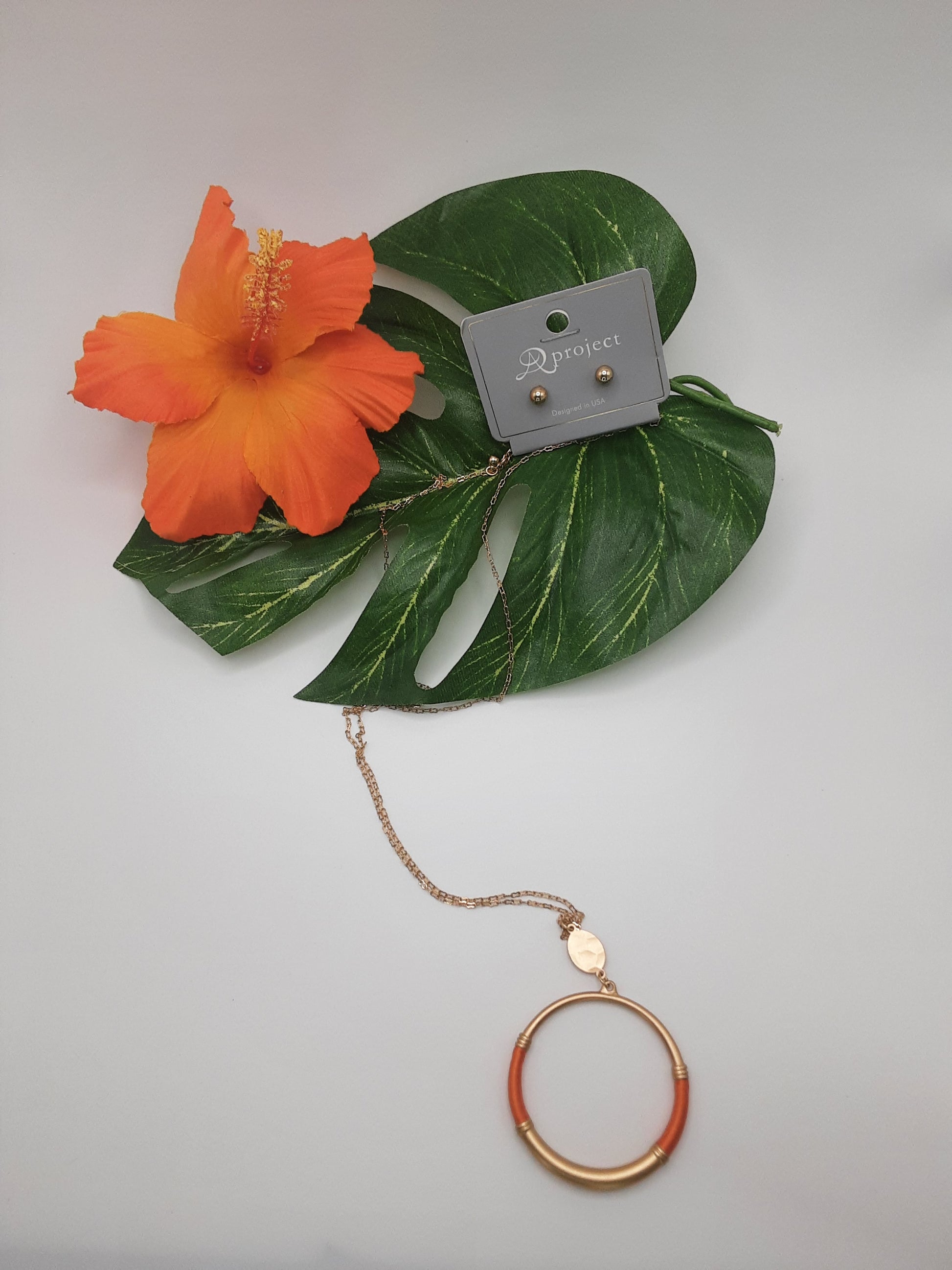 A set of APROJECT QNE3474 CIRCLE earrings from SPECIAL EFFECTS on a display card rests on green leaves with an orange hibiscus flower, and a matching orange and gold APROJECT QNE3474 CIRCLE necklace is placed below.