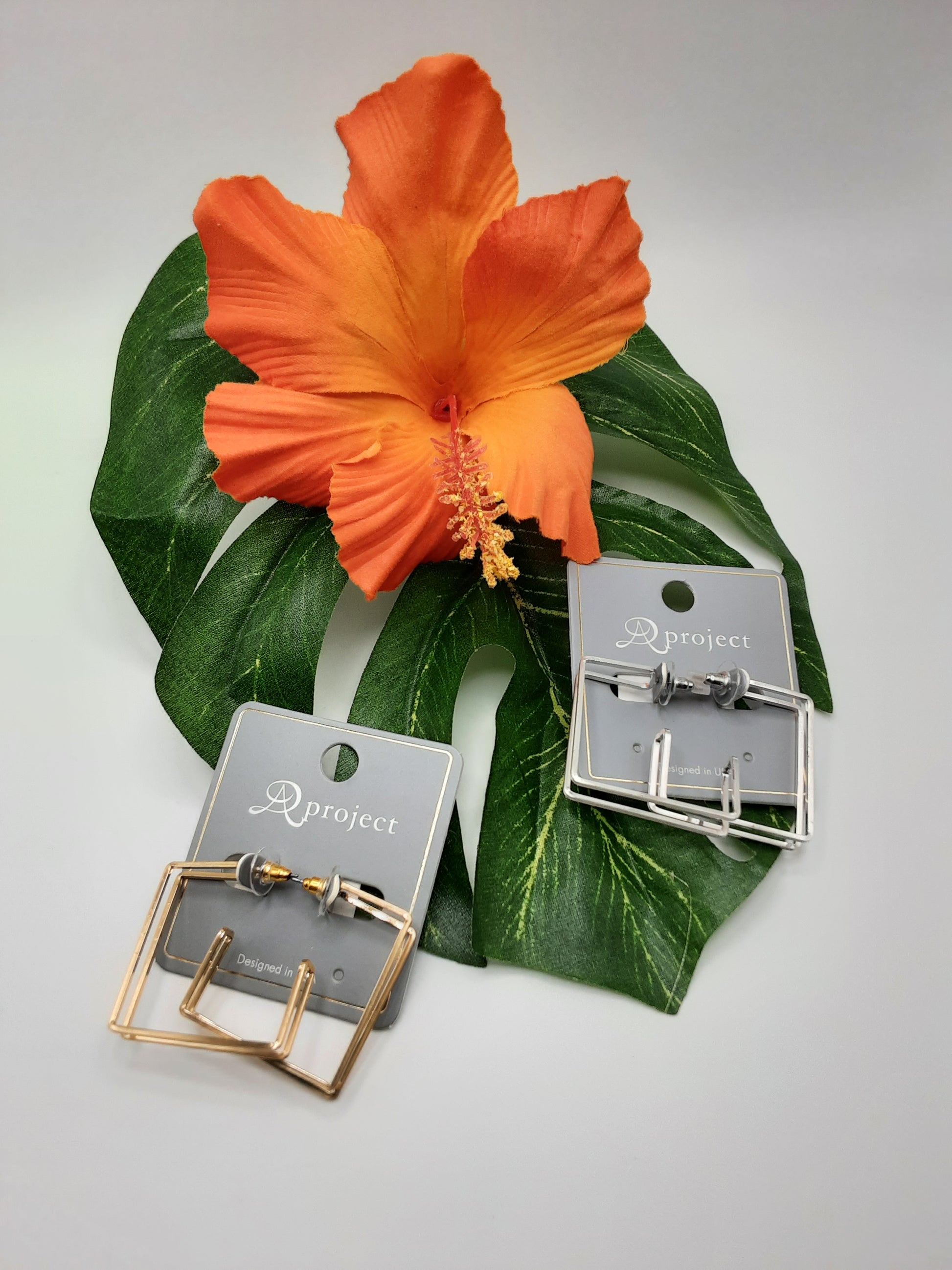 Two pairs of A PROJECT QE3243 SQUARE earrings by SPECIAL EFFECTS, featuring rectangular hoops in gold and silver, displayed on gray cards with an orange hibiscus flower and green leaves in the background.