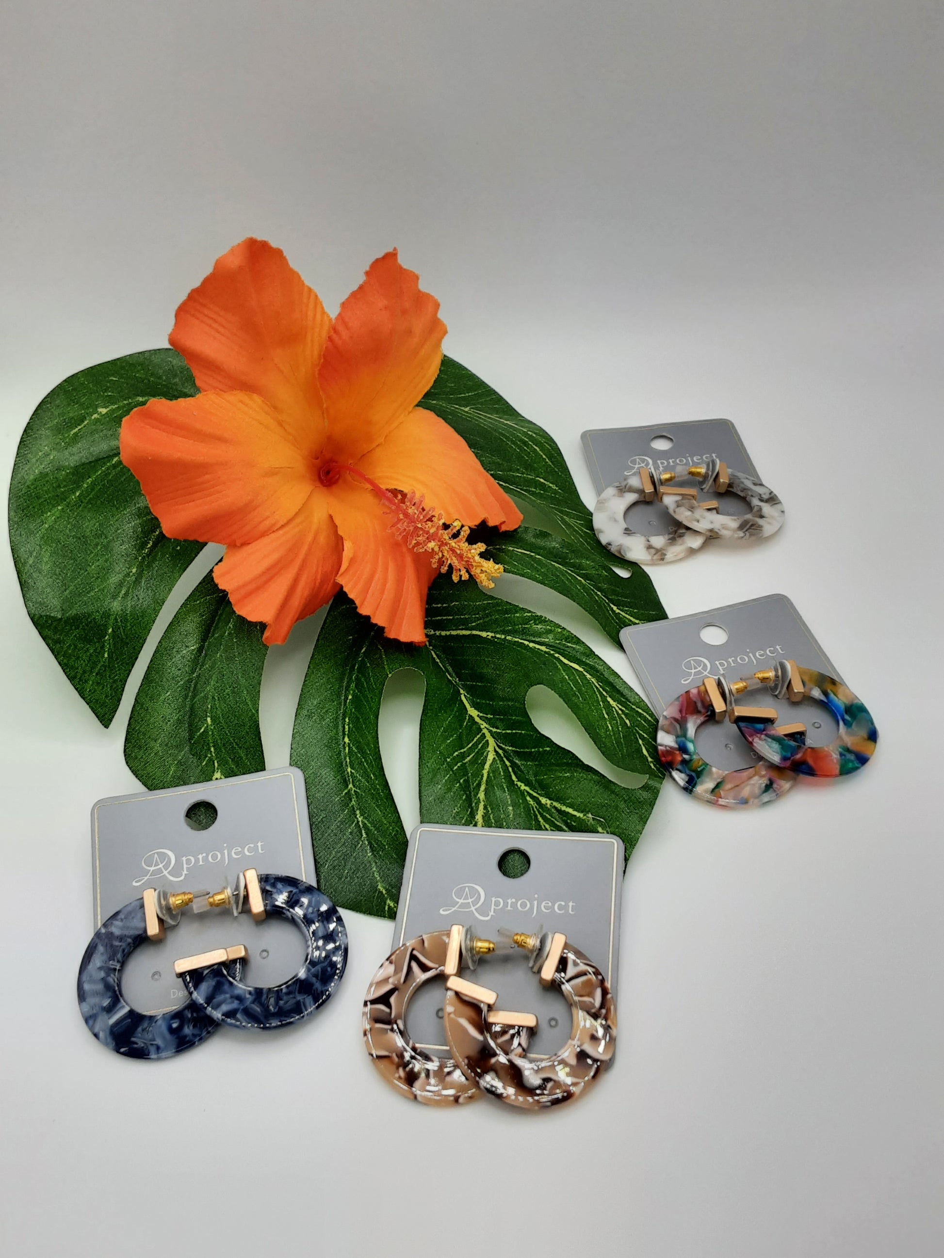 Four pairs of APROJECT QE3319 ACRYLIC hoop earrings by SPECIAL EFFECTS displayed on a tropical leaf, with a prominent orange flower in the background.