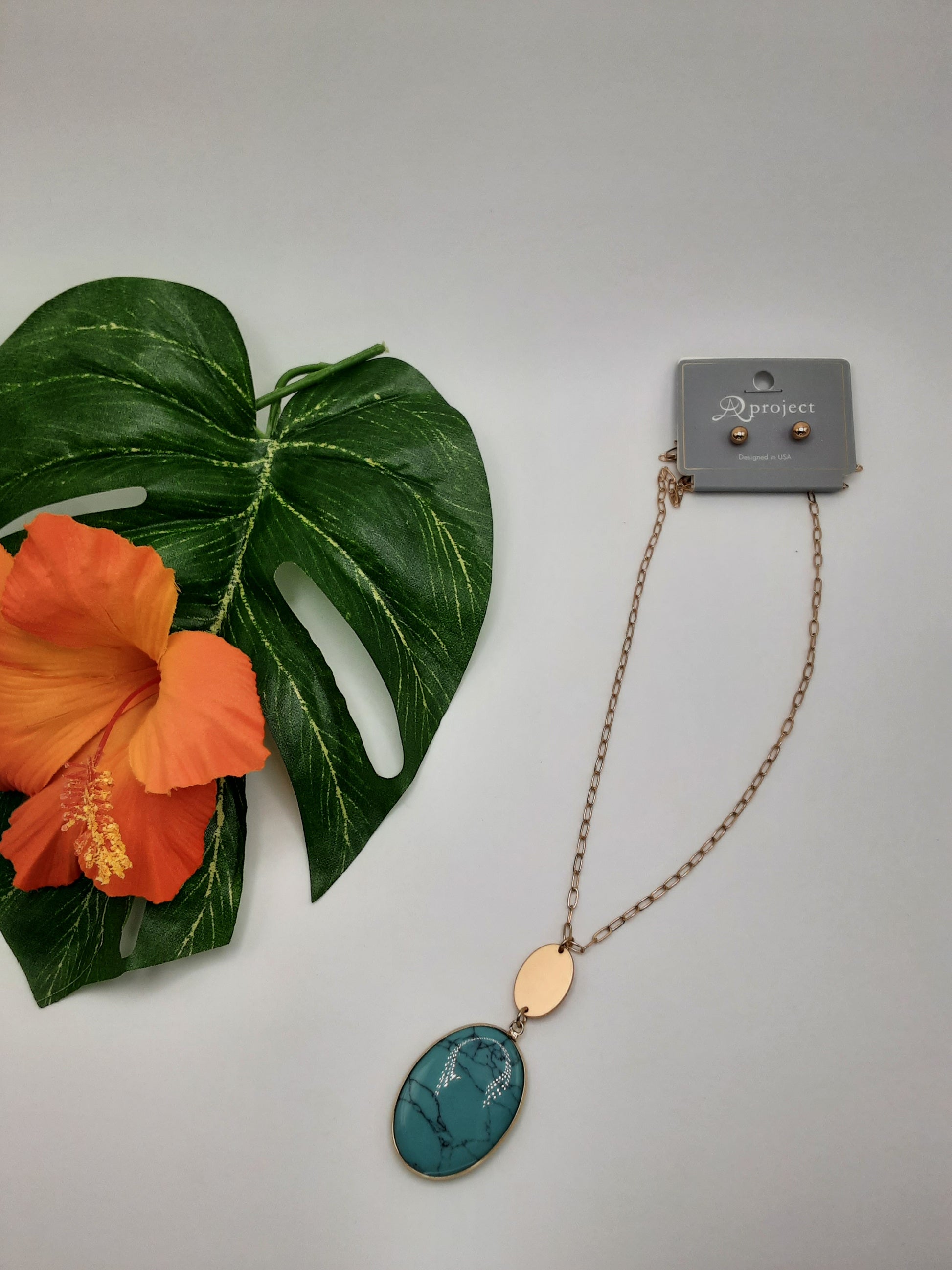 The SPECIAL EFFECTS APROJECT QNE 2342 OVAL necklace, featuring a turquoise pendant and matching earrings on a jewelry card, is displayed next to an orange hibiscus flower and a green leaf.