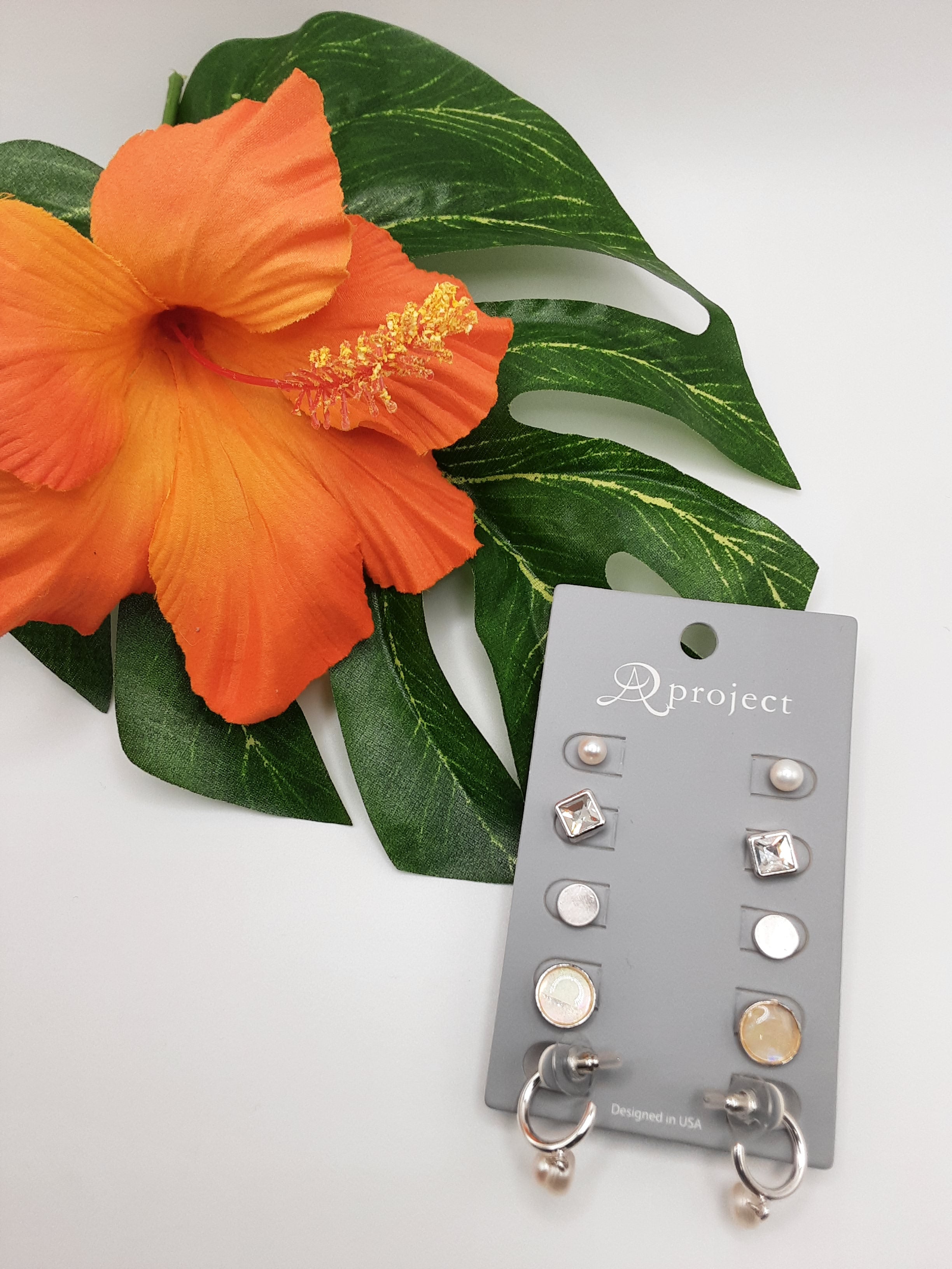 A pair of orange hibiscus flowers beside a card displaying nine assorted pairs of silver earrings. The card features the product name "A PROJECT 3596 MULTI" from the brand "SPECIAL EFFECTS. Main image