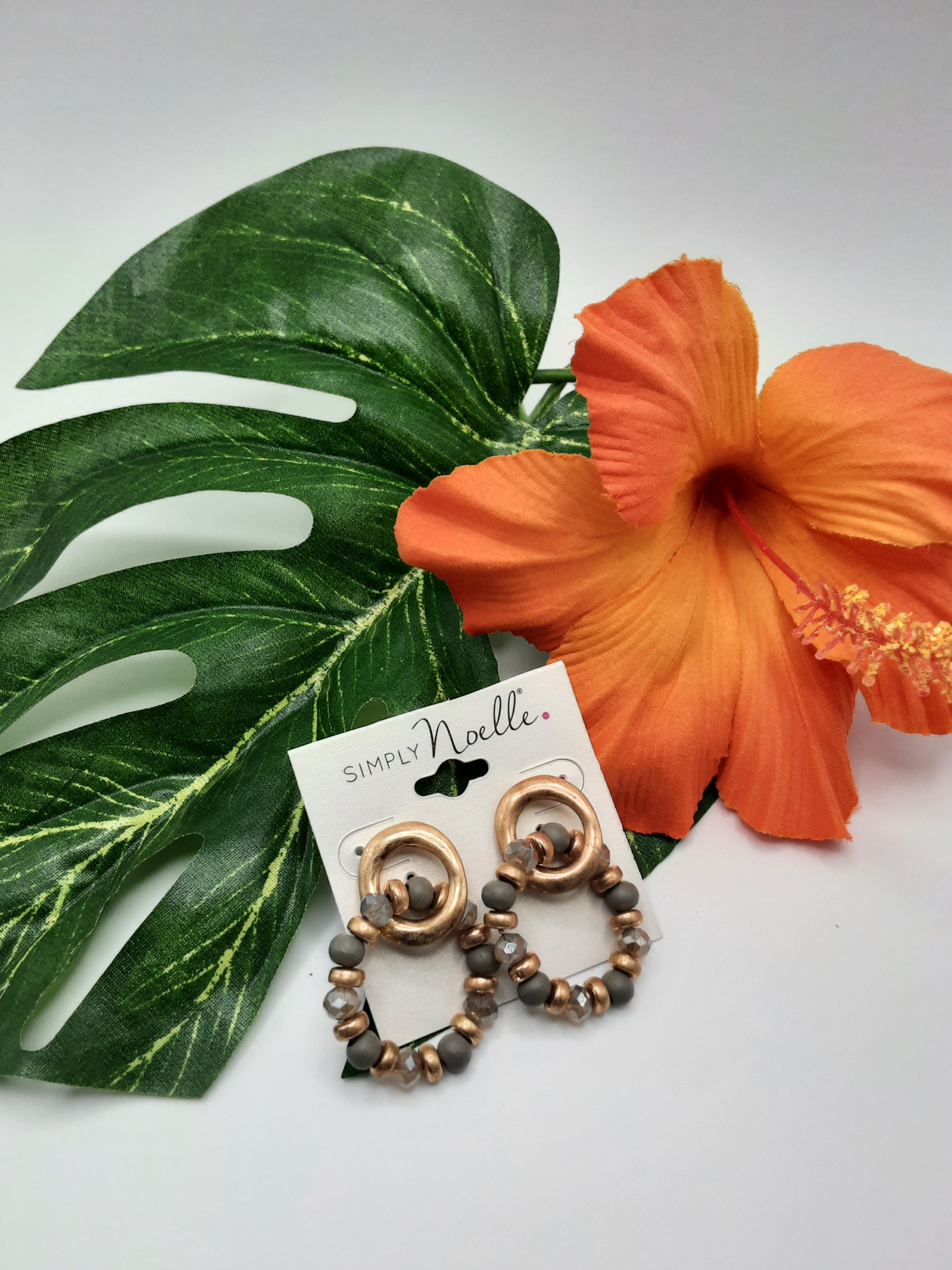 A pair of SIMPLY NOELLE SEAR102 ASST beaded hoop earrings from Ganz Inc. laid on a large green leaf next to an orange hibiscus flower. Main image