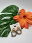 A pair of SIMPLY NOELLE SEAR102 ASST beaded hoop earrings from Ganz Inc. laid on a large green leaf next to an orange hibiscus flower.