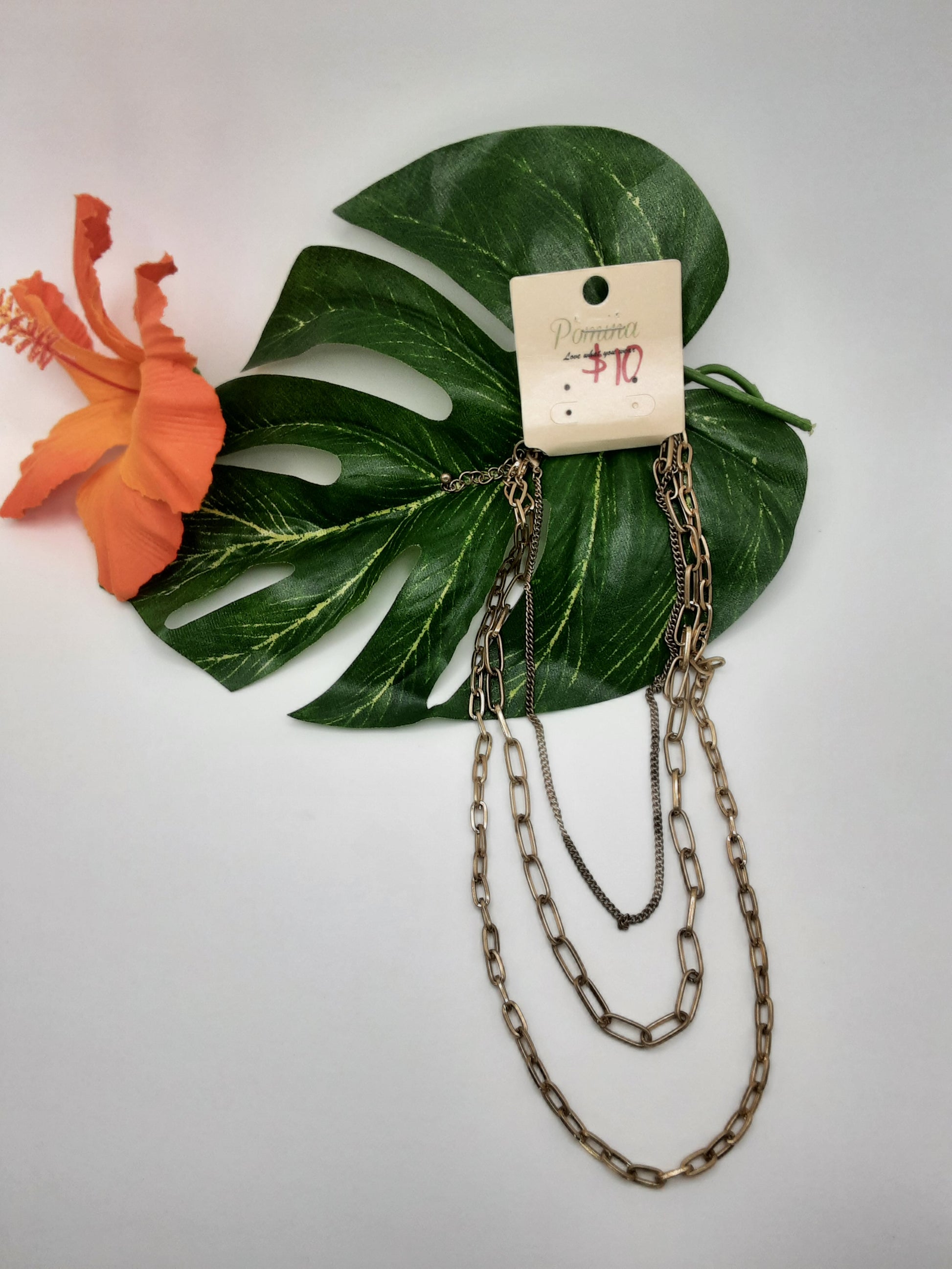 The POMINA 11843 GOLD by SPECIAL EFFECTS is a layered chain necklace, priced at $10, adorned with a large green leaf and an orange hibiscus flower.