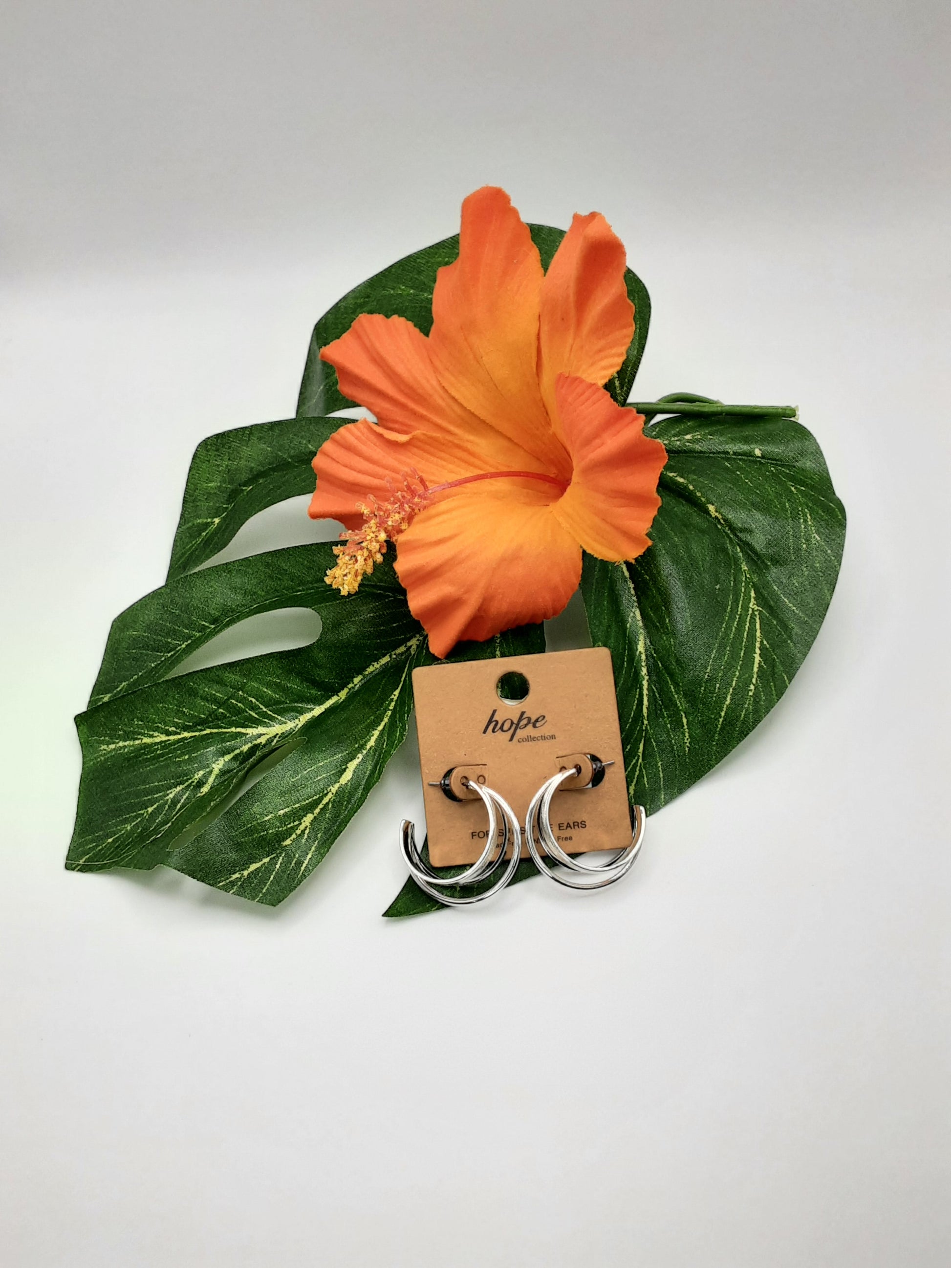 A pair of HOPE 22026 HOOP earrings by SPECIAL EFFECTS displayed on a cardboard backing labeled "Hope," set on large green leaves with an orange hibiscus flower.