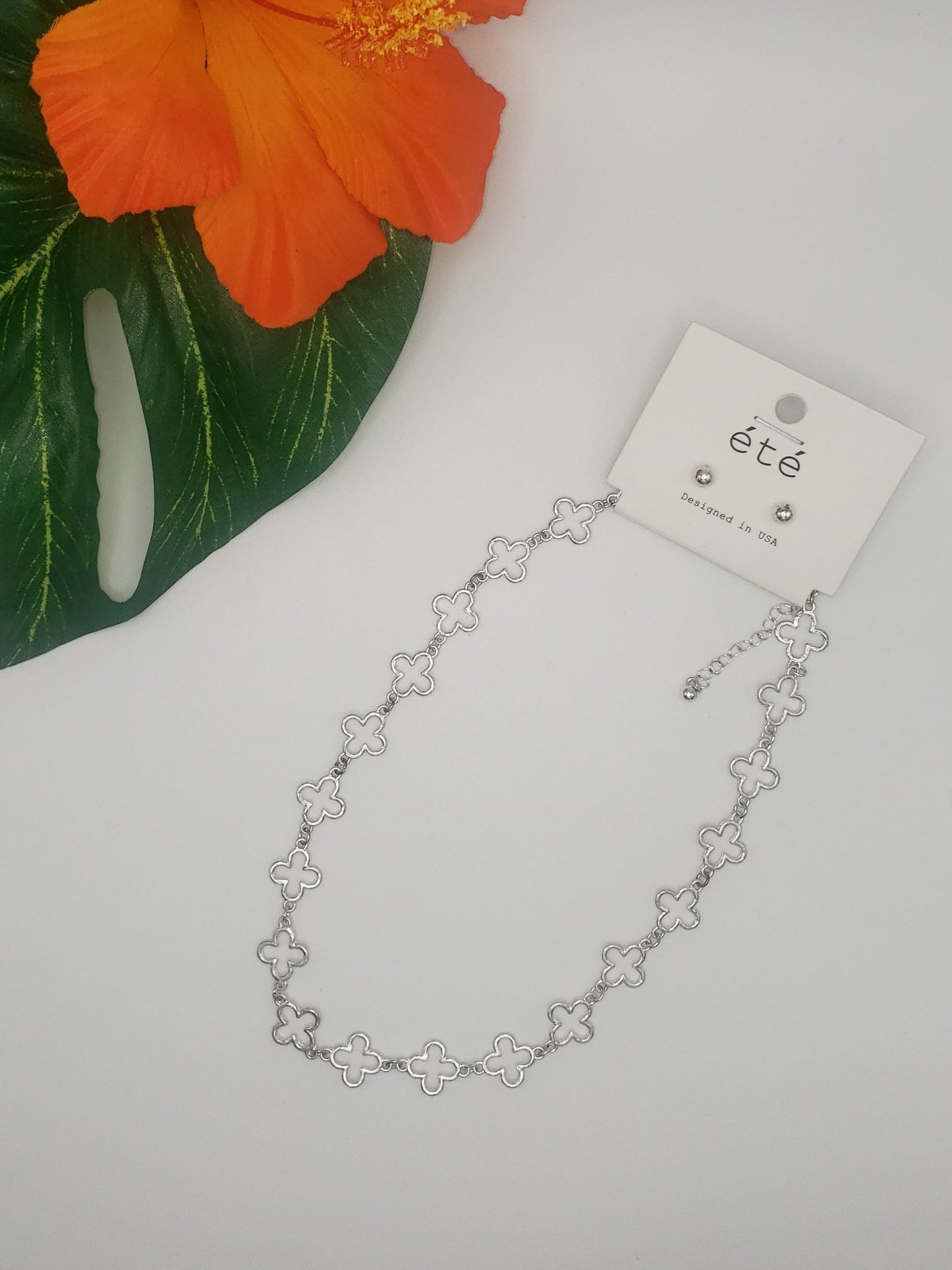 A beautiful 16-inch Clover Shaped Station Necklace from SPECIAL EFFECTS, showcasing a silver-colored clover design with a delicate floral pattern, is elegantly displayed on a white background next to a green leaf and an orange flower. The packaging features the words "été" and "TOUCH SILVER.