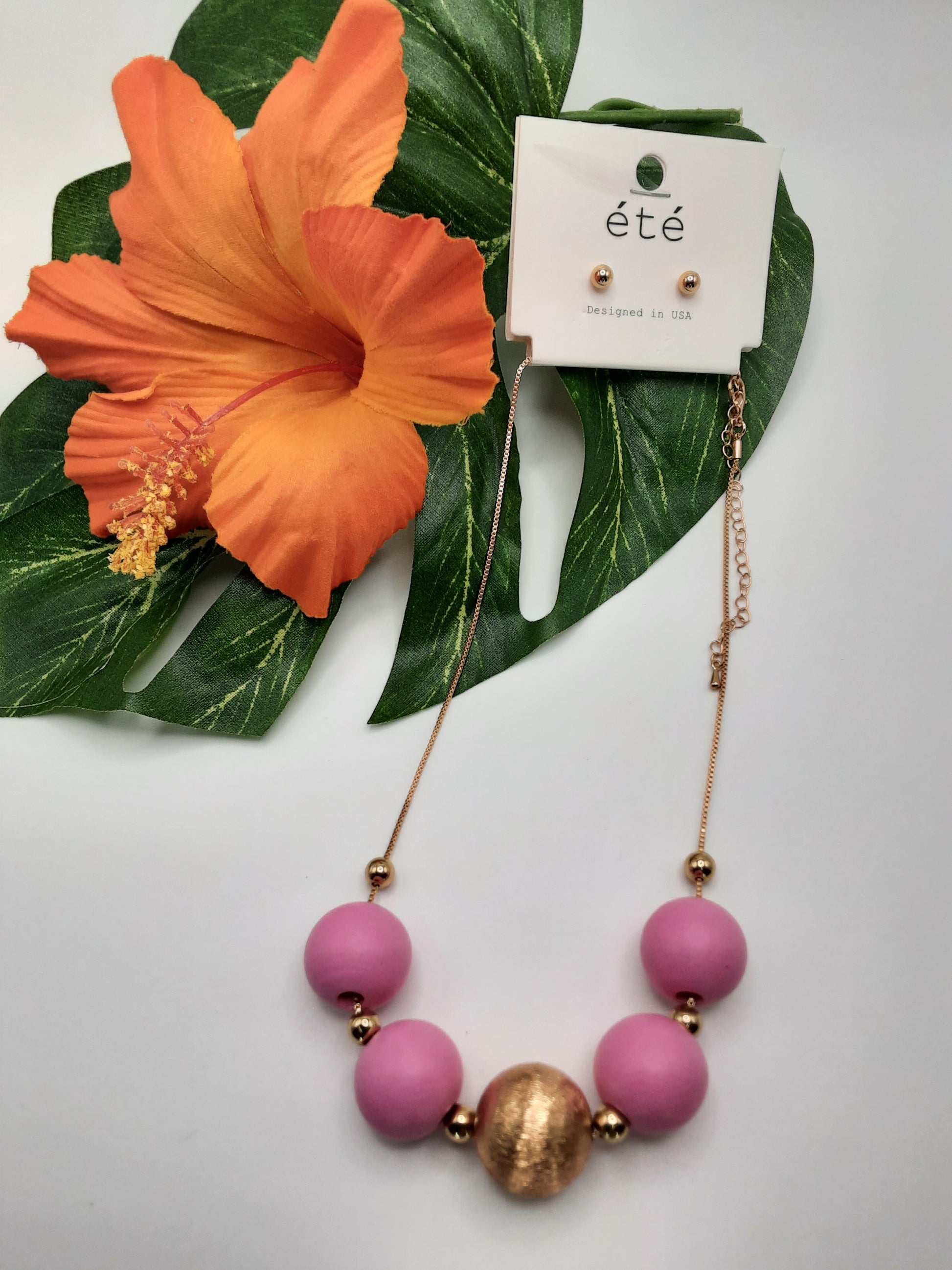 The SPECIAL EFFECTS 17" Wood Bead Necklace, featuring pastel wood beads in gold and pink, comes with matching stud earrings and is elegantly displayed on a leaf alongside an orange flower.