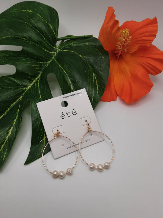 A pair of SPECIAL EFFECTS "Wire Hoops & Pearl Earrings in Silver or Gold," featuring three freshwater pearls each, are displayed on a white card labeled "été," placed beside a large green leaf and an orange hibiscus flower.