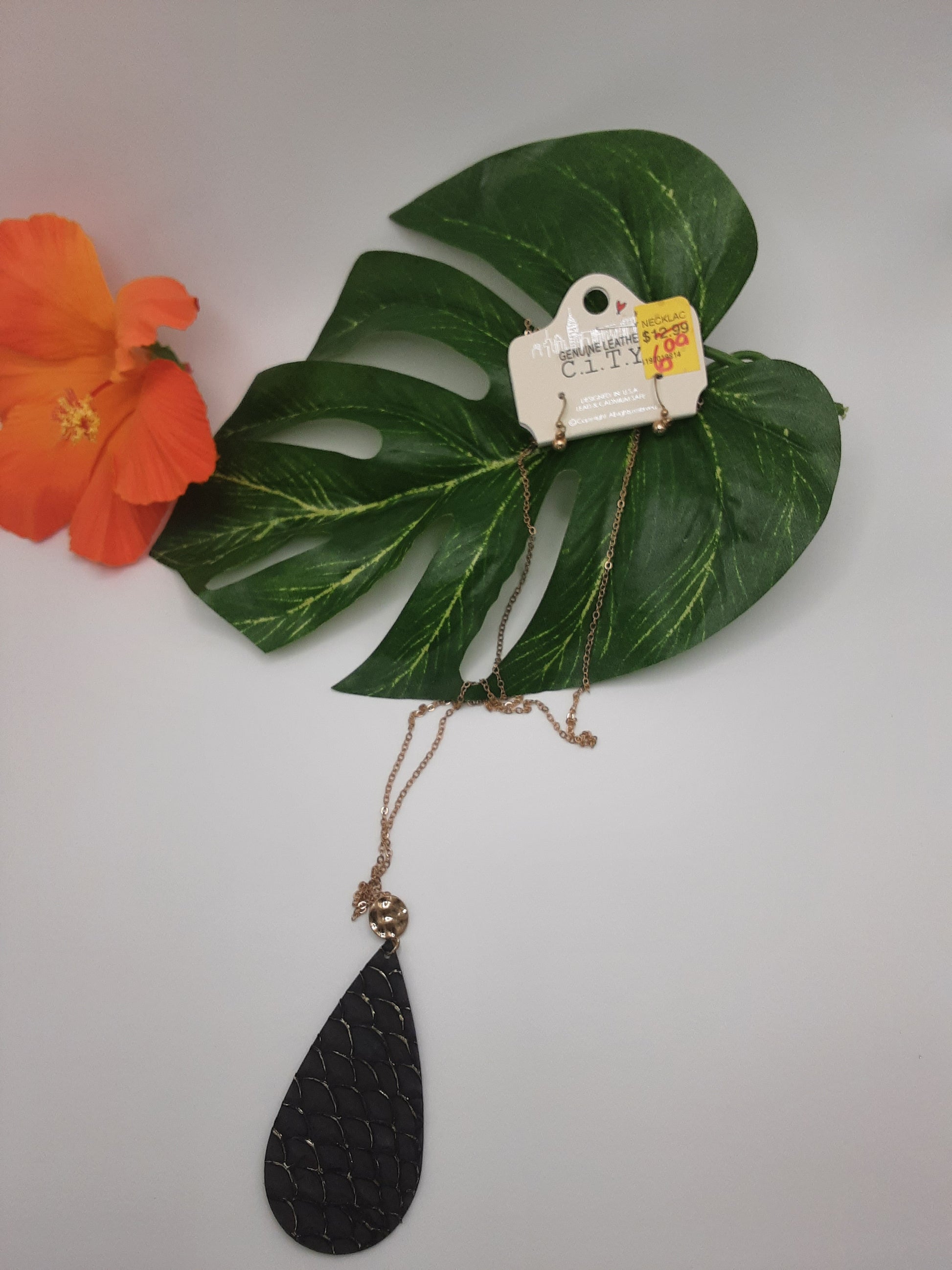 A CITY I3MNE6724 ASSORTED teardrop-shaped pendant necklace with a black pendant and gold chain from Fashion Go is displayed on a green leaf next to an orange flower. A price tag labeled "Clearance" is attached to the necklace.