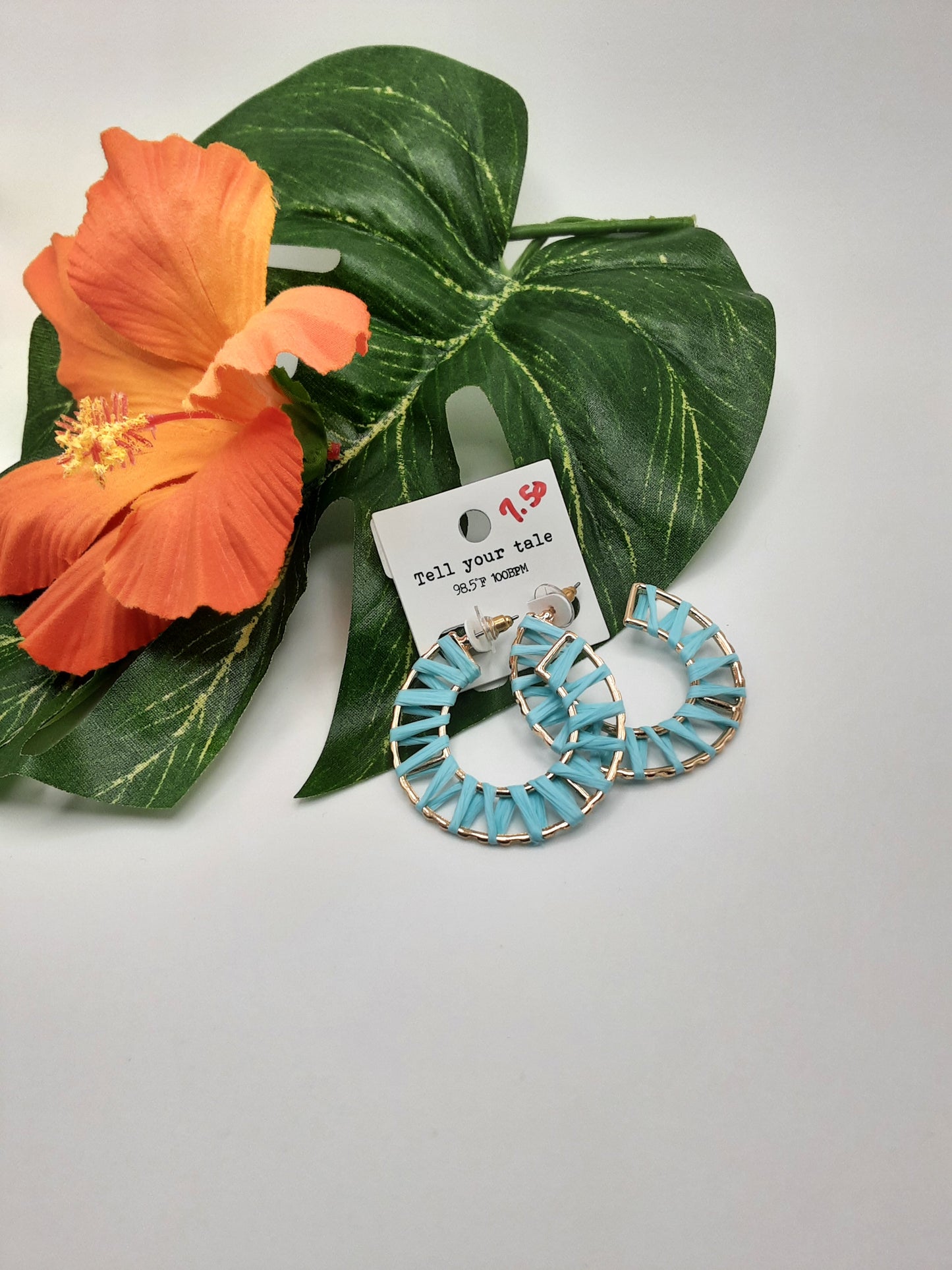 A pair of Rattan Wrapped Gold Hoop Earrings in turquoise and gold, from the brand SPECIAL EFFECTS, displayed on a green leaf with an orange flower. Accompanied by a tag that reads "Tell your tale" at the top.