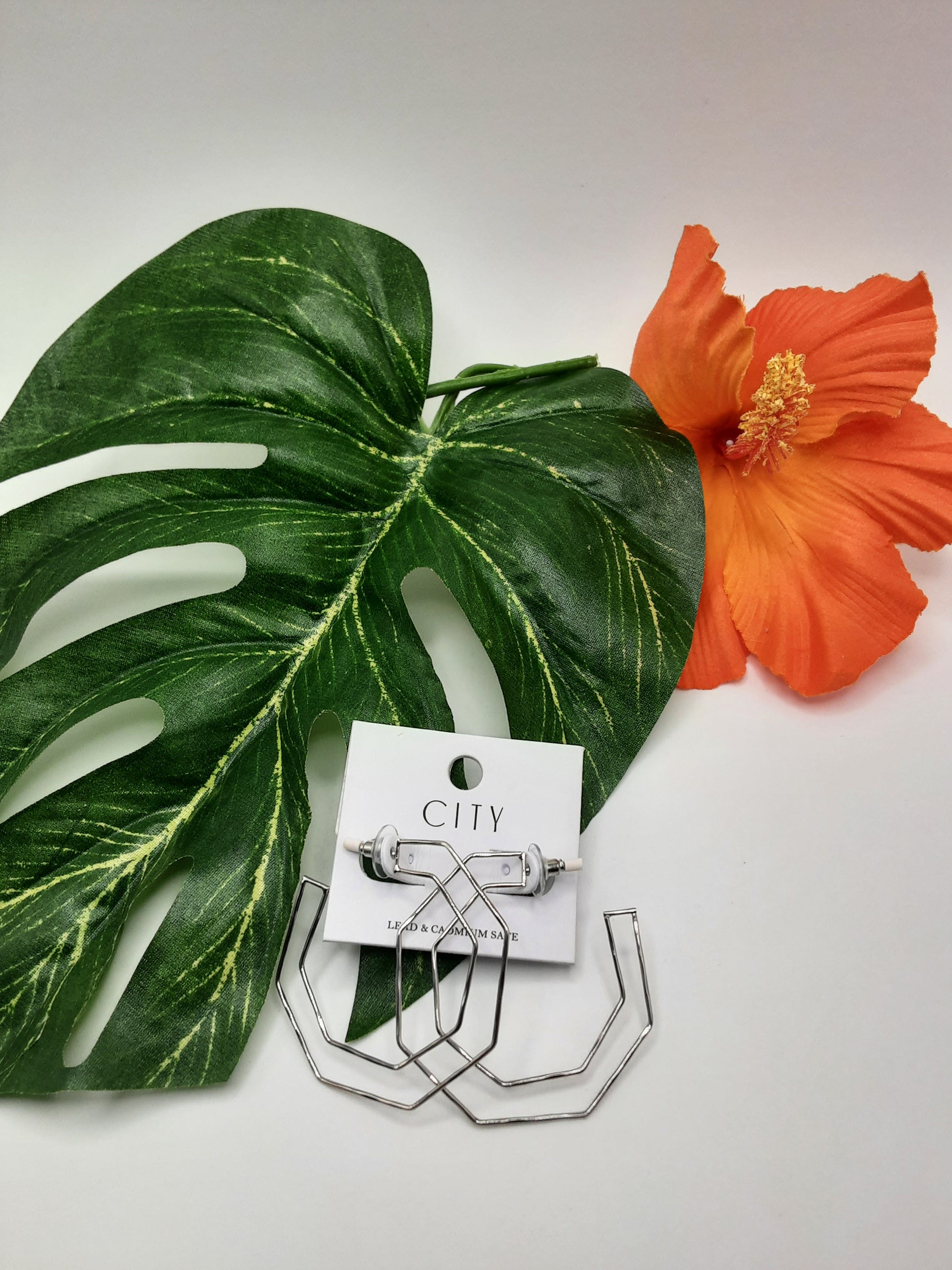 The CITY BE1675RH OCTAGON silver geometric earrings by SPECIAL EFFECTS are beautifully displayed on a white card, elegantly arranged on a green monstera leaf with an orange flower beside them.