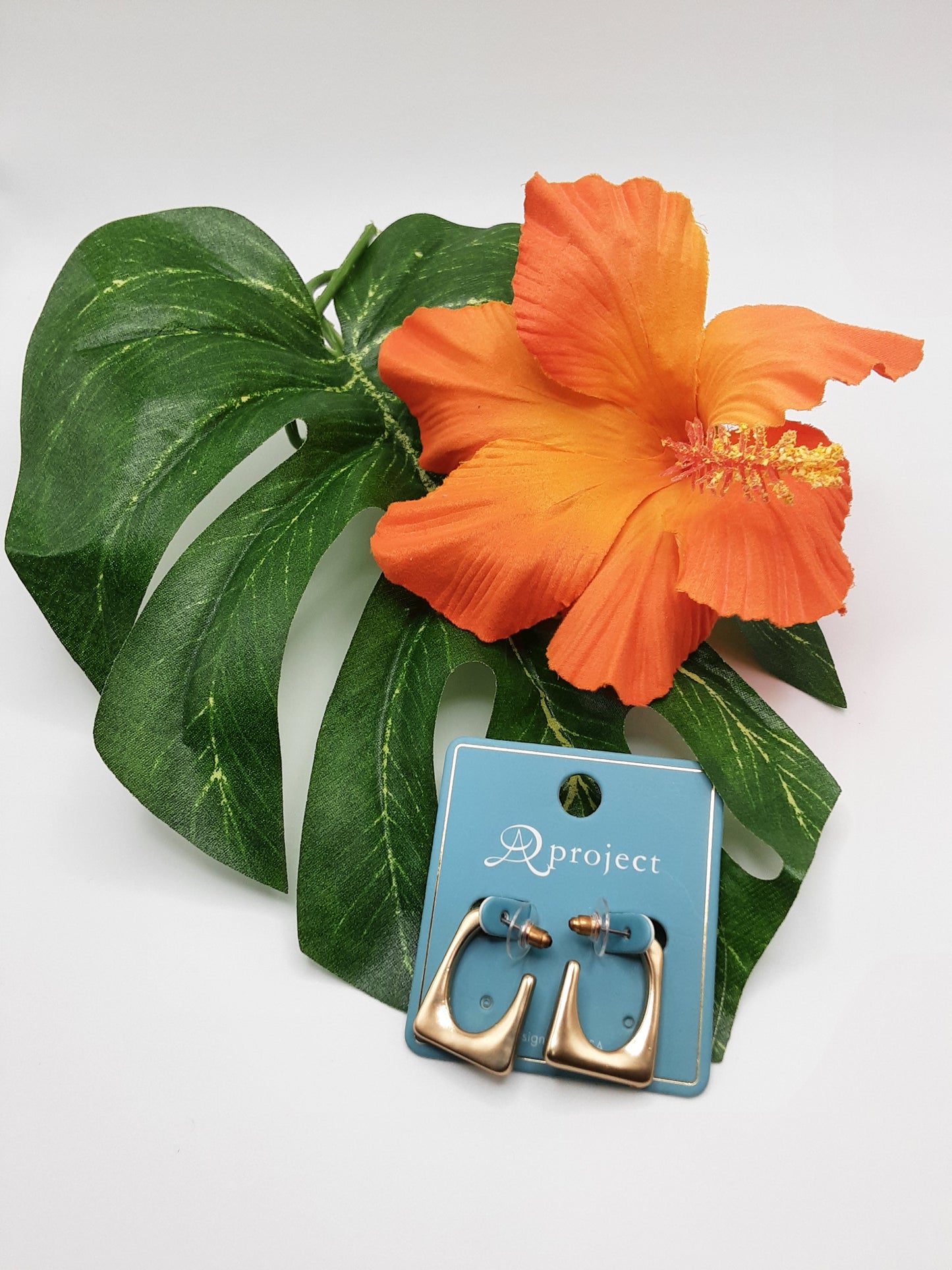 A pair of SPECIAL EFFECTS APROJECT QE2739 ASST gold and turquoise earrings displayed on a blue card with "Dproject" text, placed beside a green leaf and an orange flower on a white background.