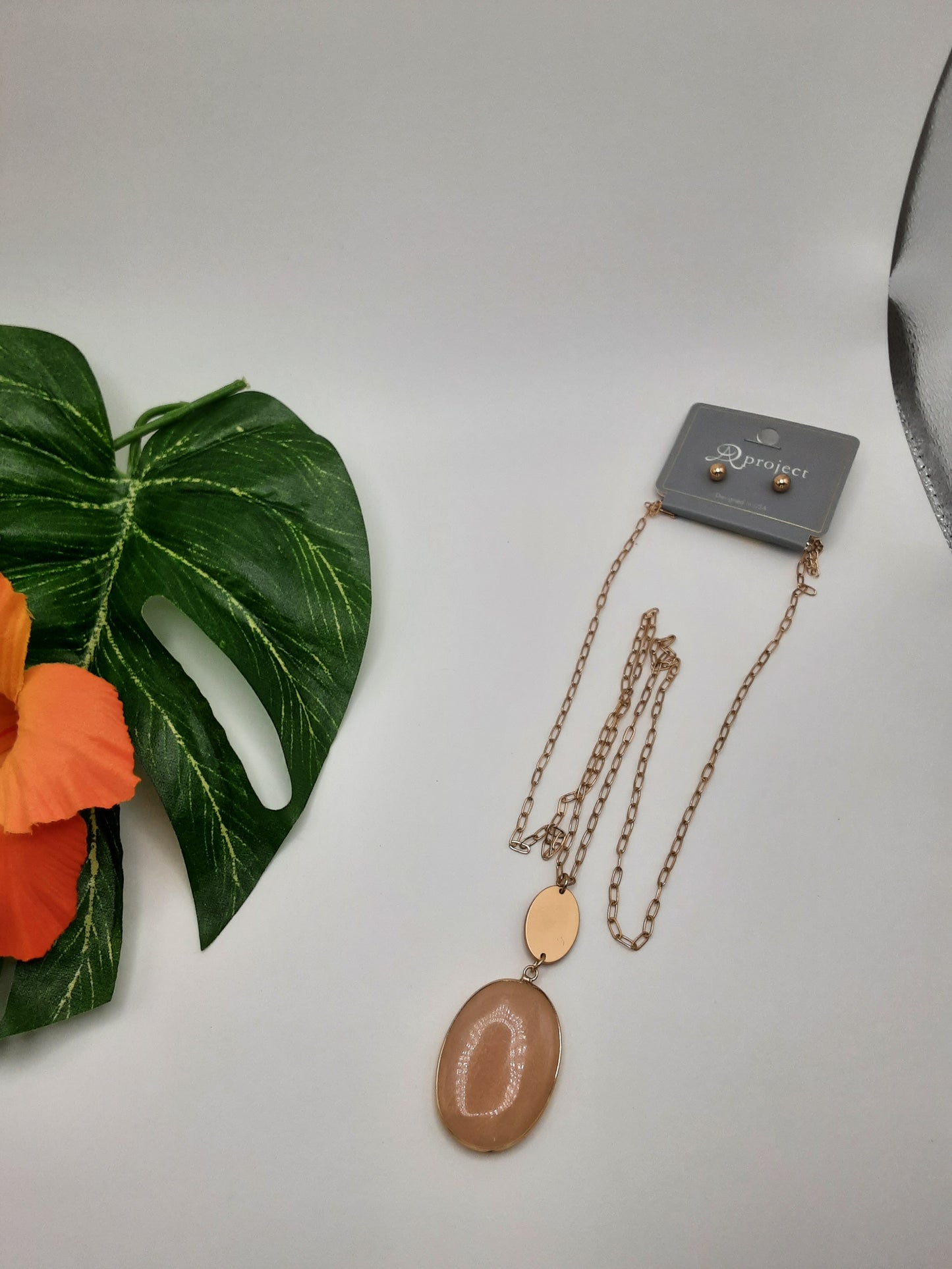 The APROJECT QNE 2342 OVAL gold chain necklace by SPECIAL EFFECTS, featuring an oval pendant, is elegantly displayed next to a green leaf and an orange flower on a white background.