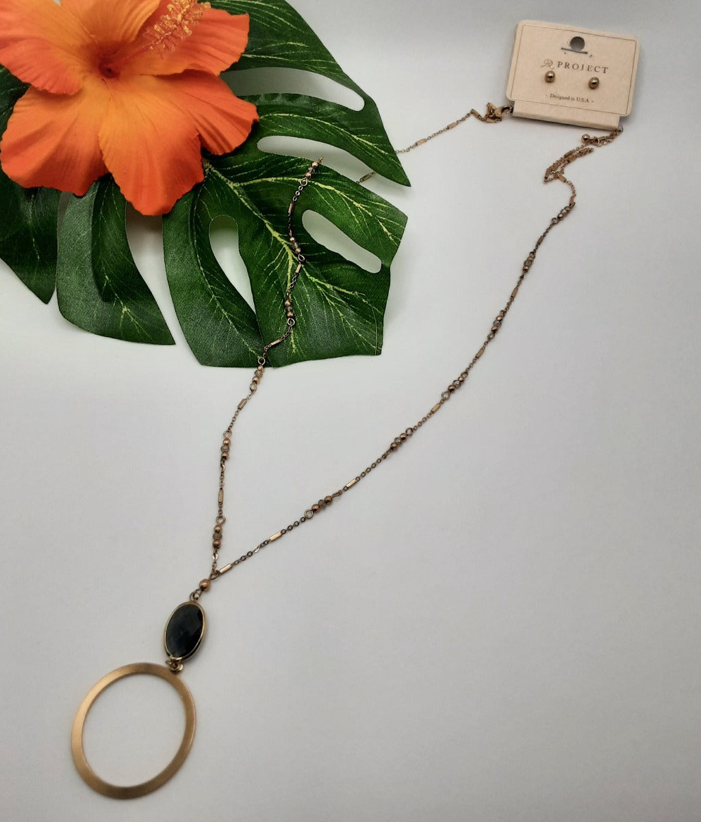 The PROJECT QNE1289 SMOKE necklace by SPECIAL EFFECTS, featuring a circular pendant adorned with a dark oval stone, is showcased alongside a vibrant orange hibiscus flower and lush green leaves.