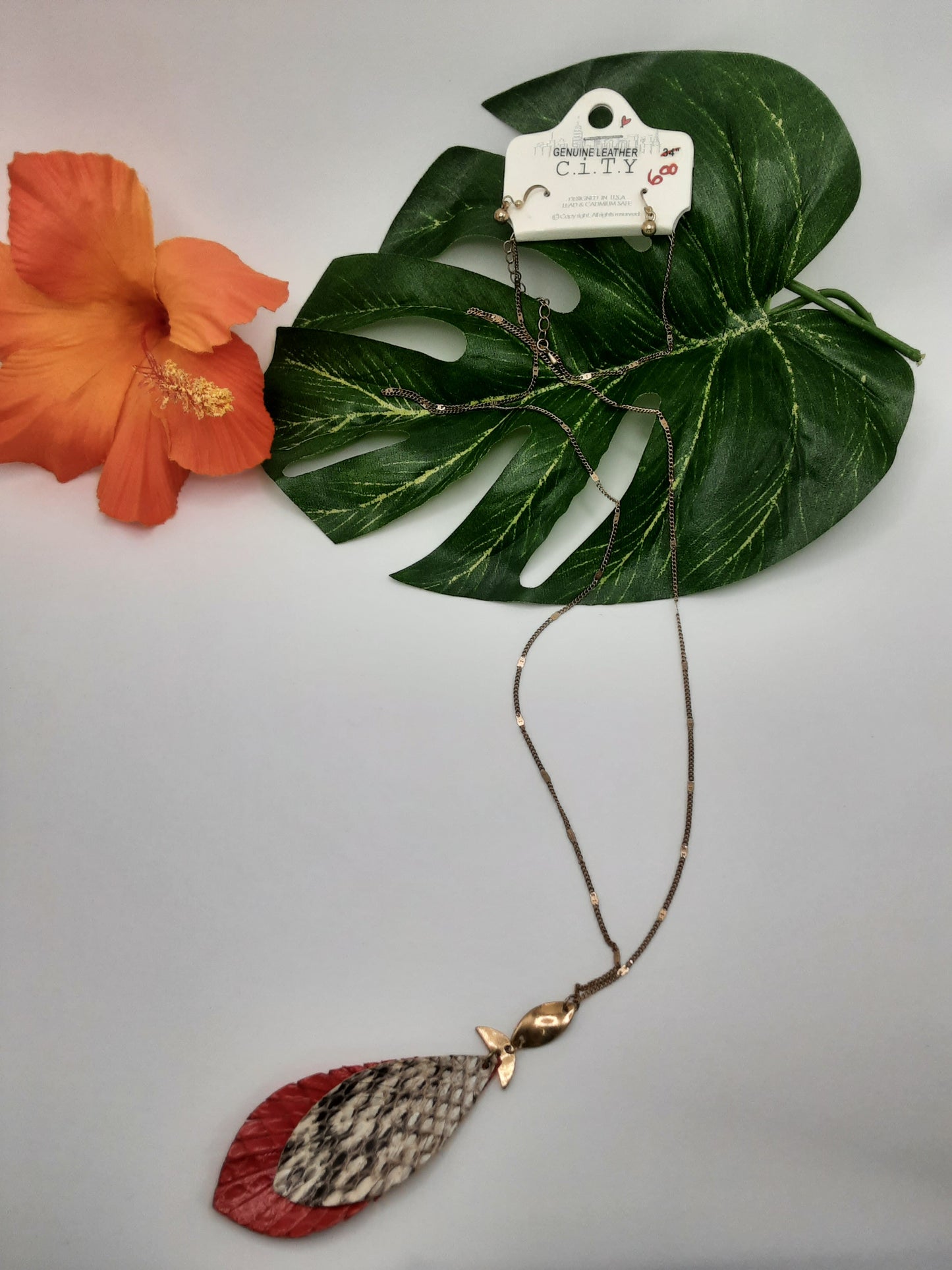 The CITY 176821 ASSORTED necklace by Fashion Go features a pendant with three feather-like elements in red, white, and black. It is displayed on a leaf with an orange flower placed to the left. The price tag reads $8.00.