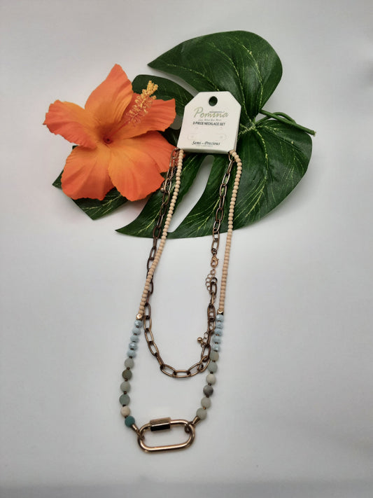 The POMINA BO1JN0576 ASSORTED beaded necklace by Fashion Go, featuring a combination of aqua and beige beads, chain links, and a large gold clasp, is showcased on a white background next to an orange hibiscus flower and green leaves.