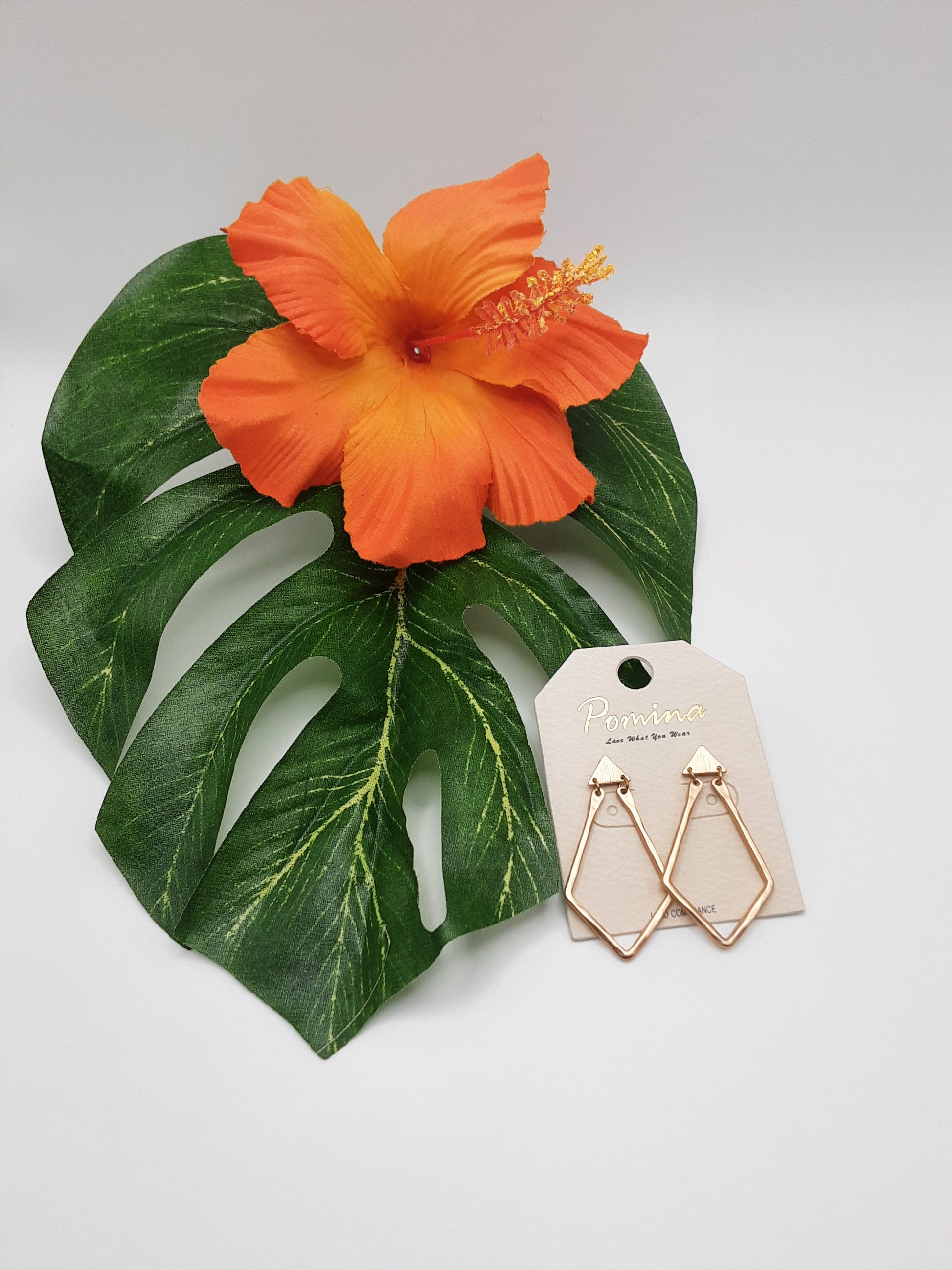 The POMINA HE3242 METAL earrings from Fashion Go are showcased on a label, resting on a large green leaf with an orange hibiscus flower.