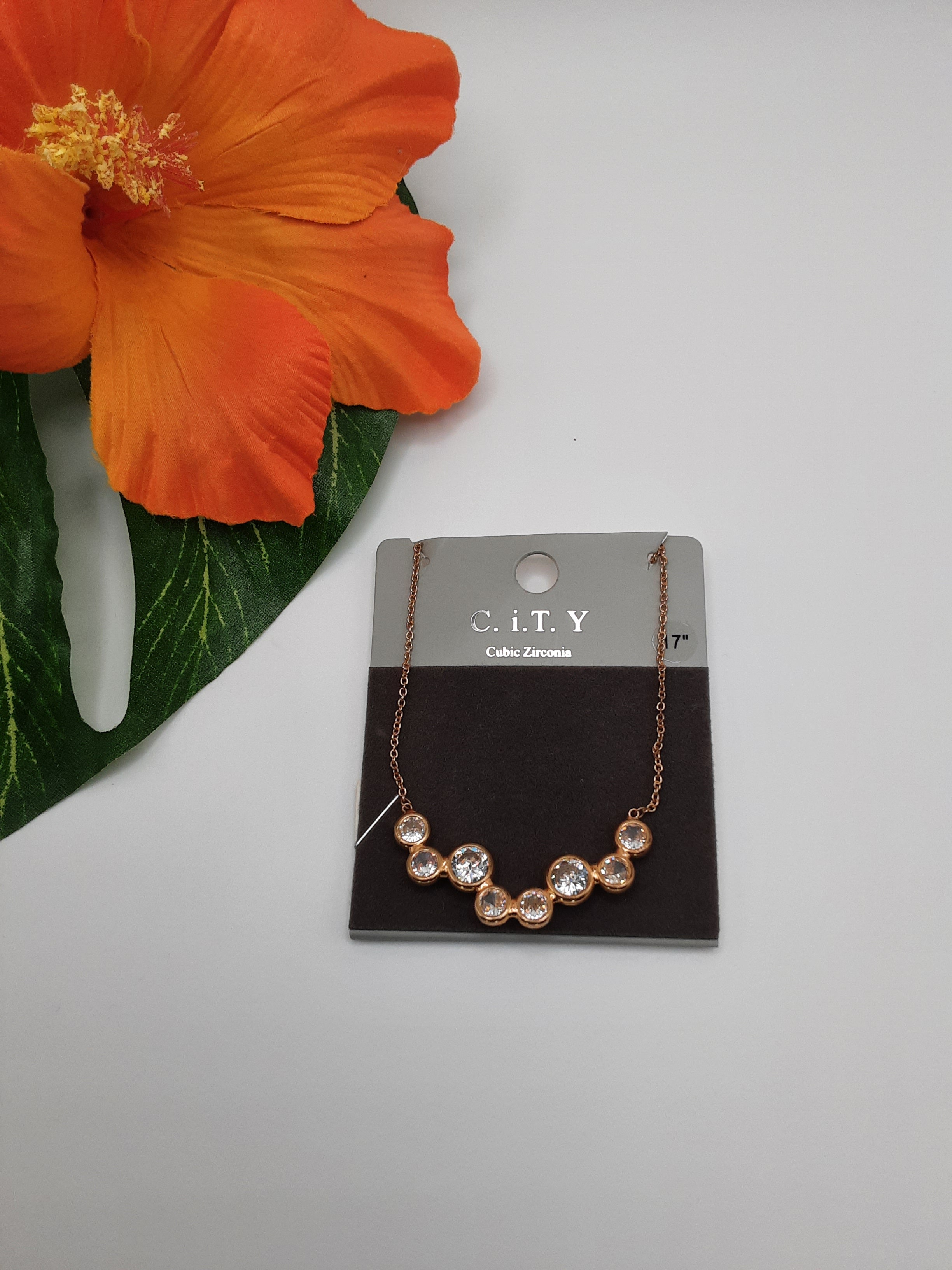A necklace with seven round cubic zirconia stones, named the CITY MNE6082 CZ by SPECIAL EFFECTS, is displayed on a black and gray card labeled "C.I.T.Y Cubic Zirconia," next to an orange flower and green leaf. Main image