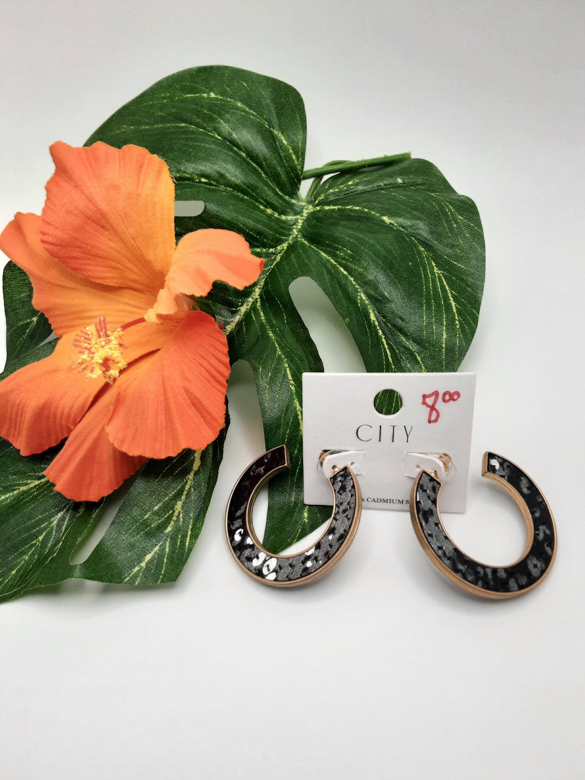 A pair of black and gold hoop earrings from the CITY ME7988 SNAKE collection by SPECIAL EFFECTS, displayed on a white card featuring "CITY" branding, placed on a green leaf accompanied by an orange flower. The price "$8.00" is handwritten in red on the card.