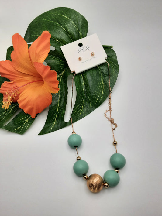 A jewelry set featuring the 17" Wood Bead Necklace by SPECIAL EFFECTS, adorned with large green and gold beads, displayed on a green leaf with an orange flower, and paired with small gold earrings on a card reading "été".