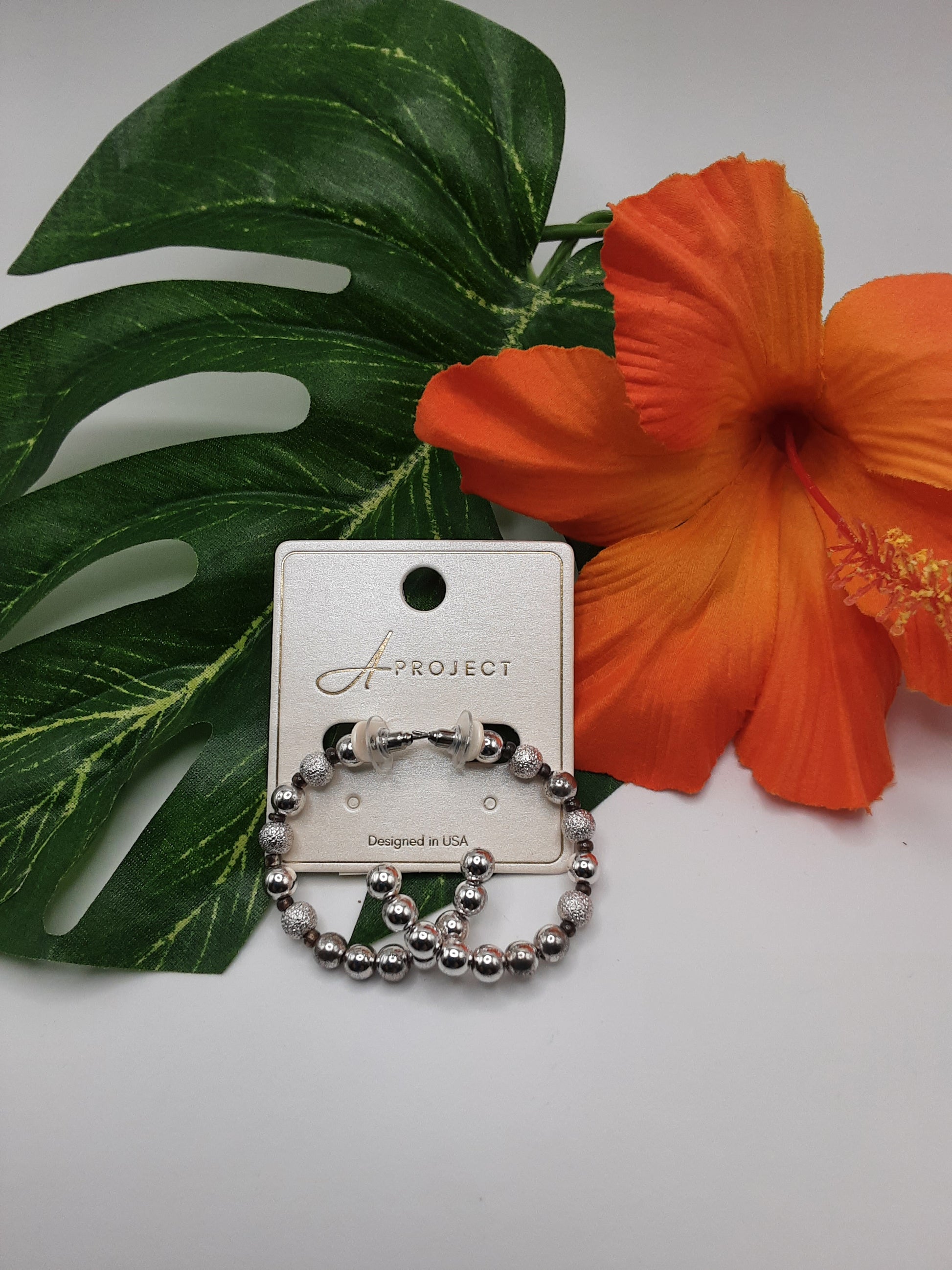 The APROJECT KE2297 HOOP earrings by SPECIAL EFFECTS are showcased on a card adorned with a green leaf and an orange flower for decoration.